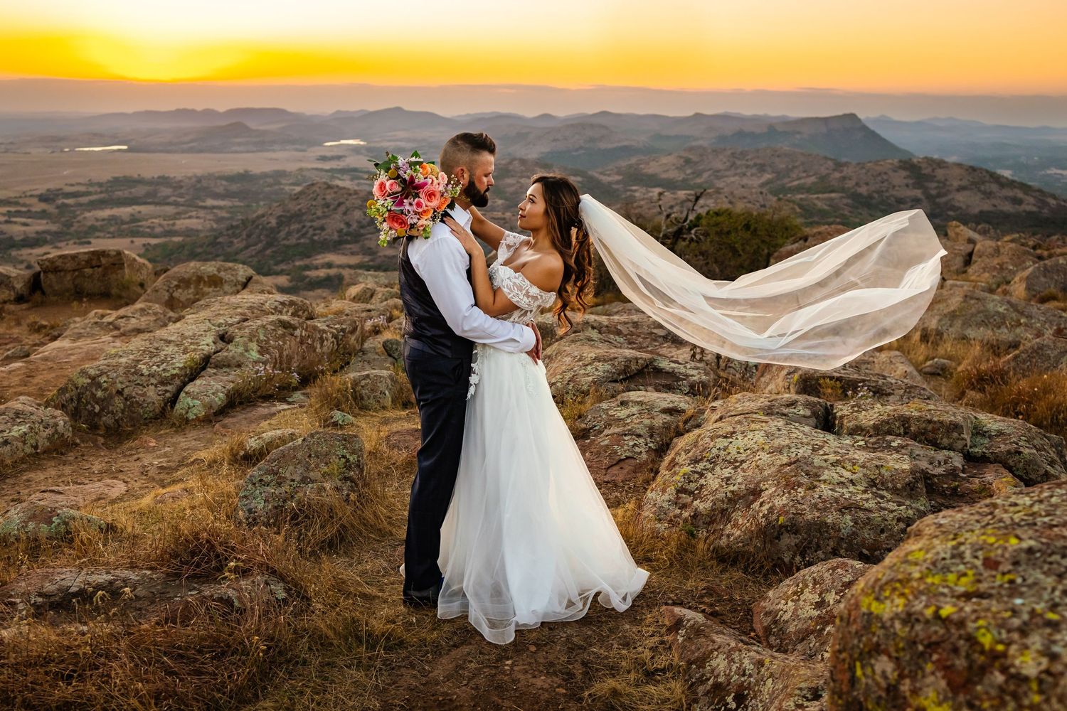 Bride and groom embrace on rocky Oklahoma Mount Scott hilltop at sunset, veil flowing in wind, with scenic valley landscape behind them.