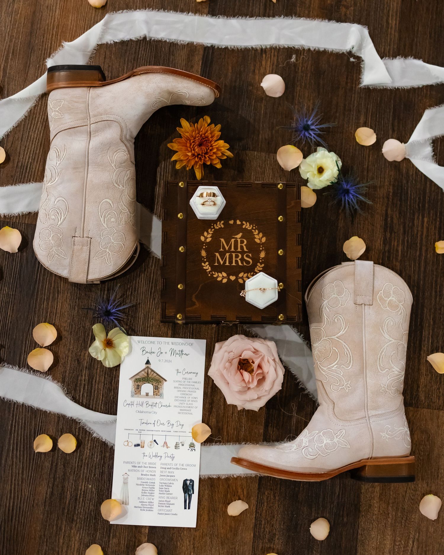White cowboy boots, wedding rings, Mr & Mrs book, and floral accents arranged on rustic wood for an Oklahoma country wedding flat lay.