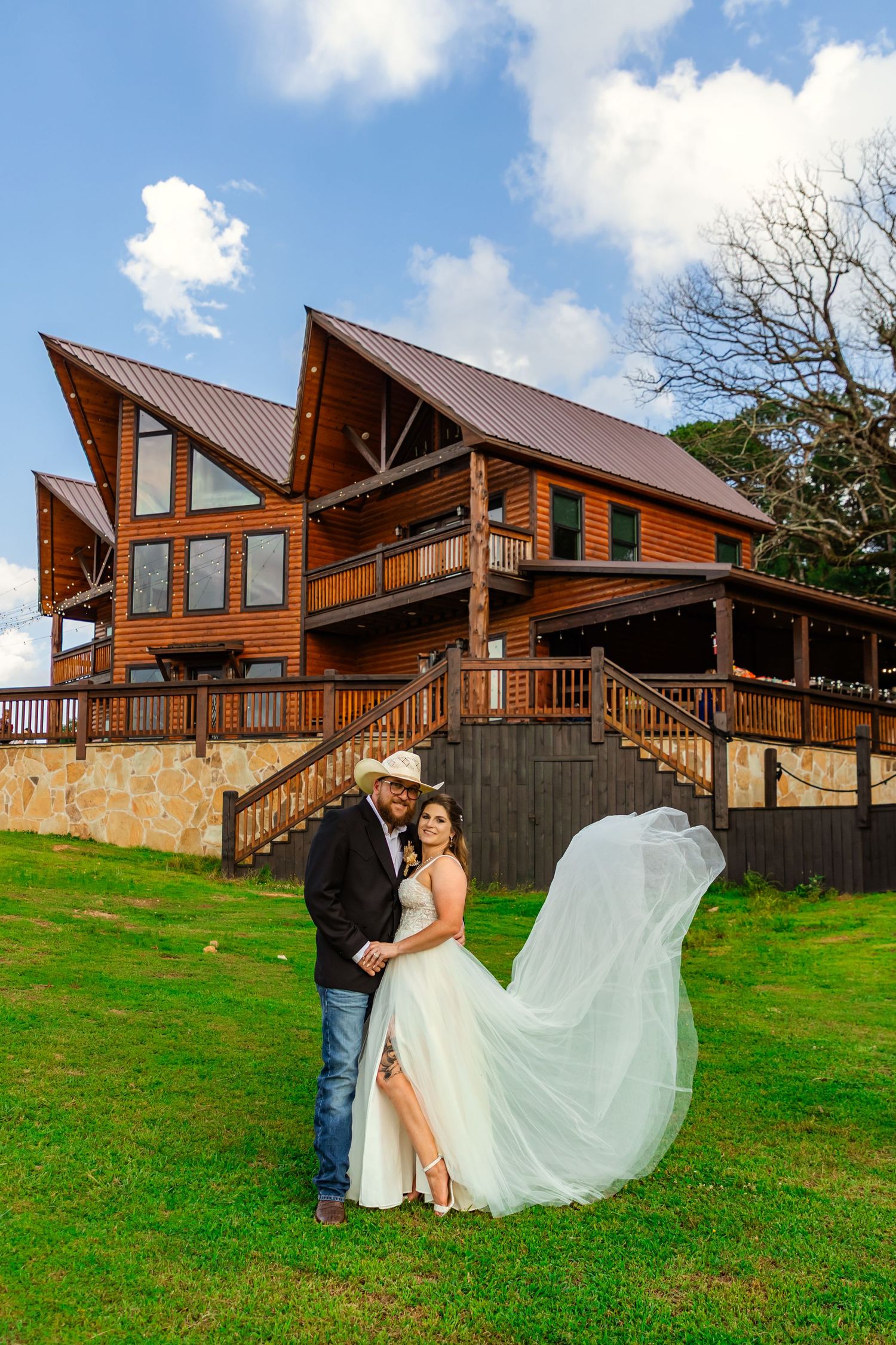 Bride and groom embrace outdoors in front of a rustic log cabin venue in broken bow, her veil flowing in the breeze under a blue sky.