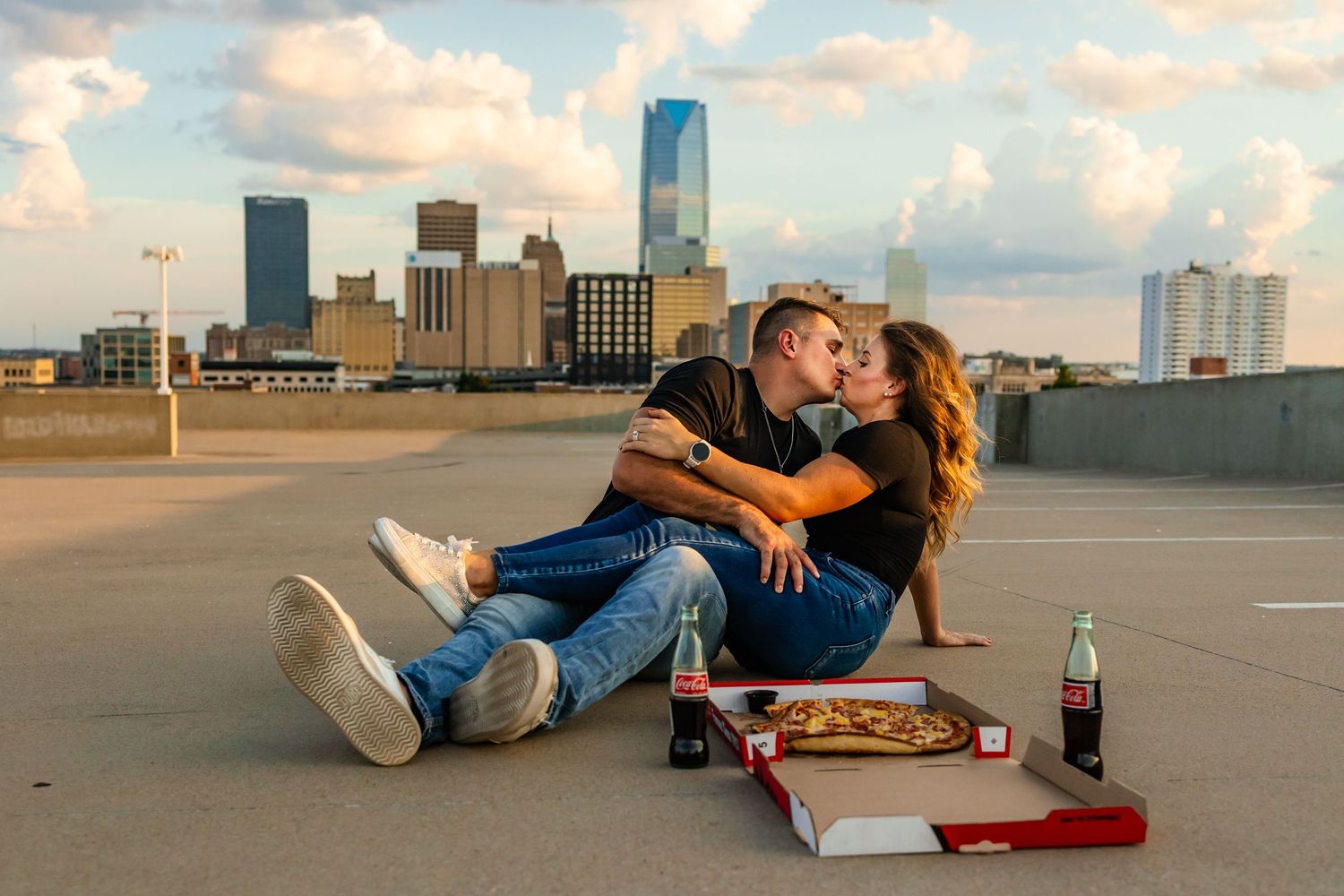 Oklahoma couple kissing on rooftop with pizza boxes and soda, city skyline in background at sunset.