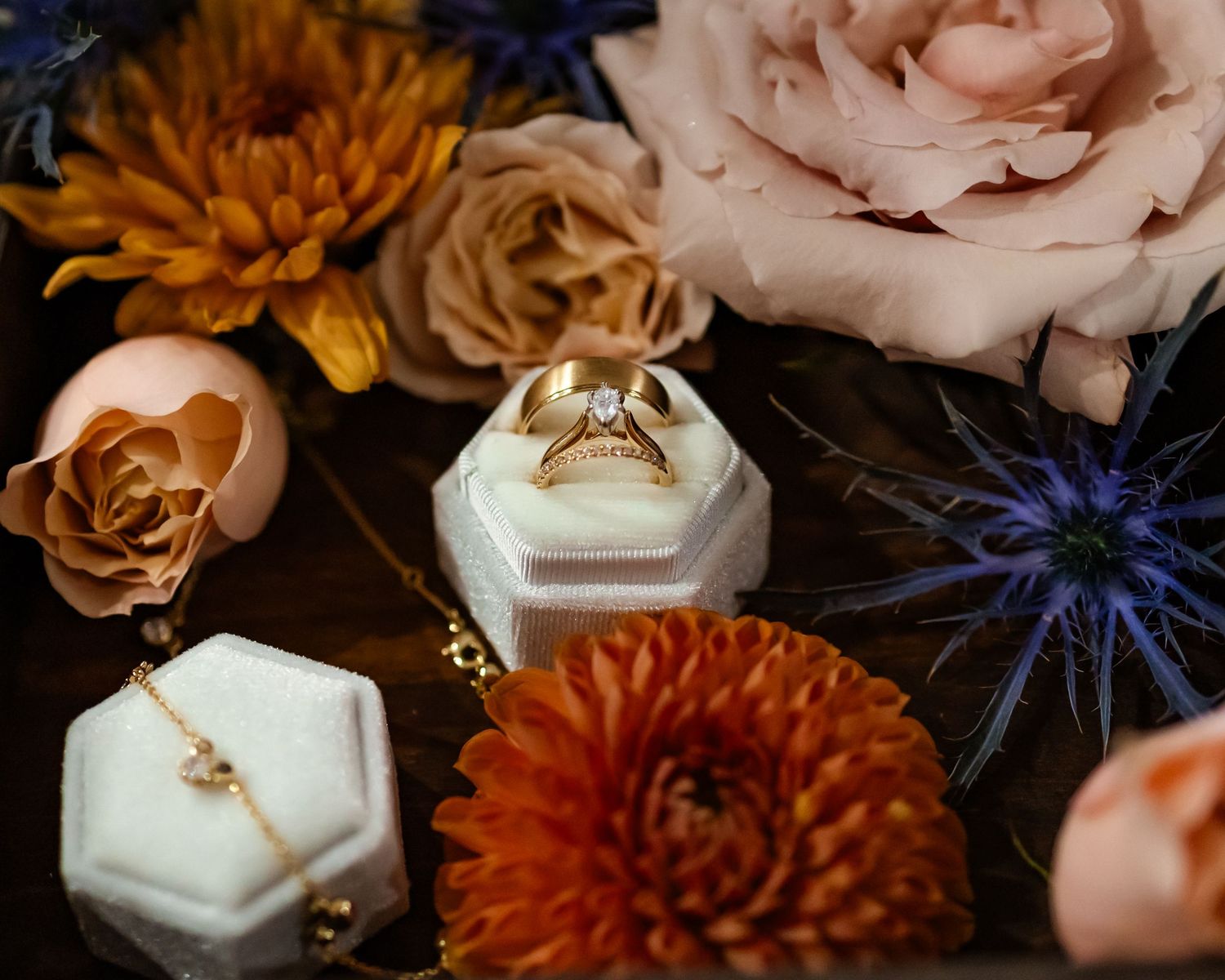 Gold diamond engagement ring displayed in a white velvet hexagon box surrounded by orange and blush roses and blue thistle flowers.