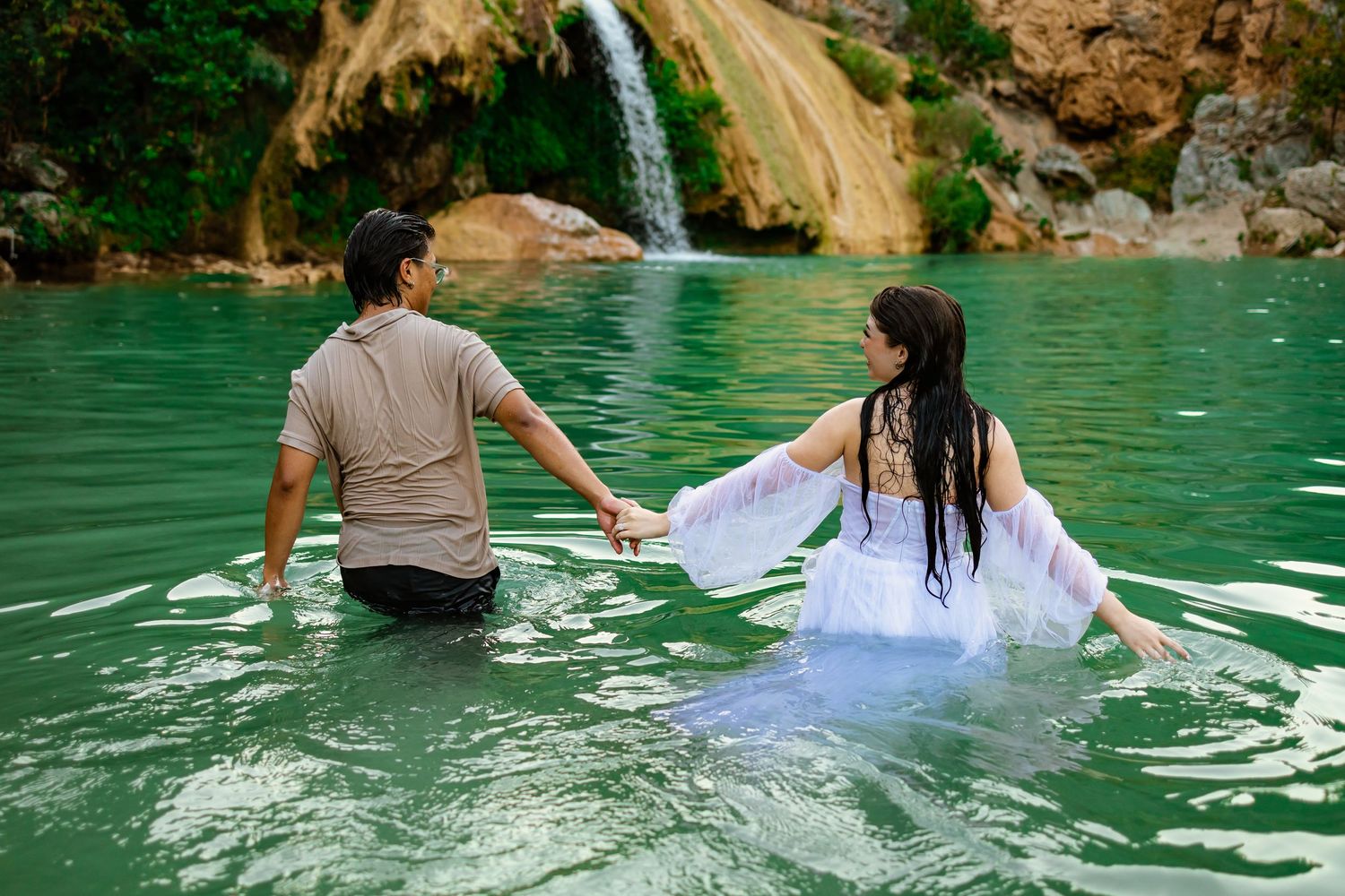 Couple holding hands wading in turquoise water near a waterfall, woman in white dress, romantic outdoor scene.