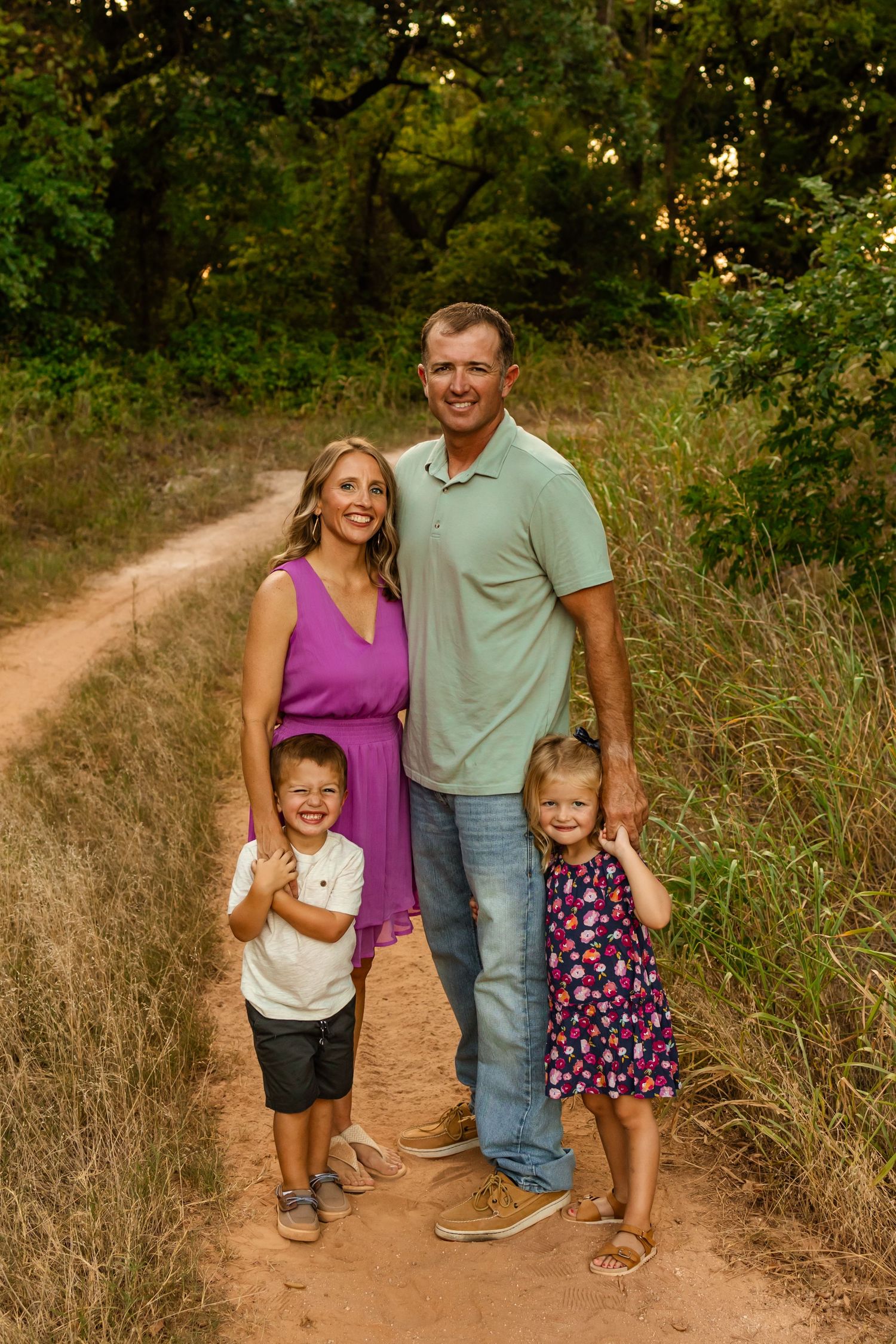 Family of four posing on an Oklahoma dirt trail surrounded by lush greenery, parents with two young children smiling outdoors. family shoot