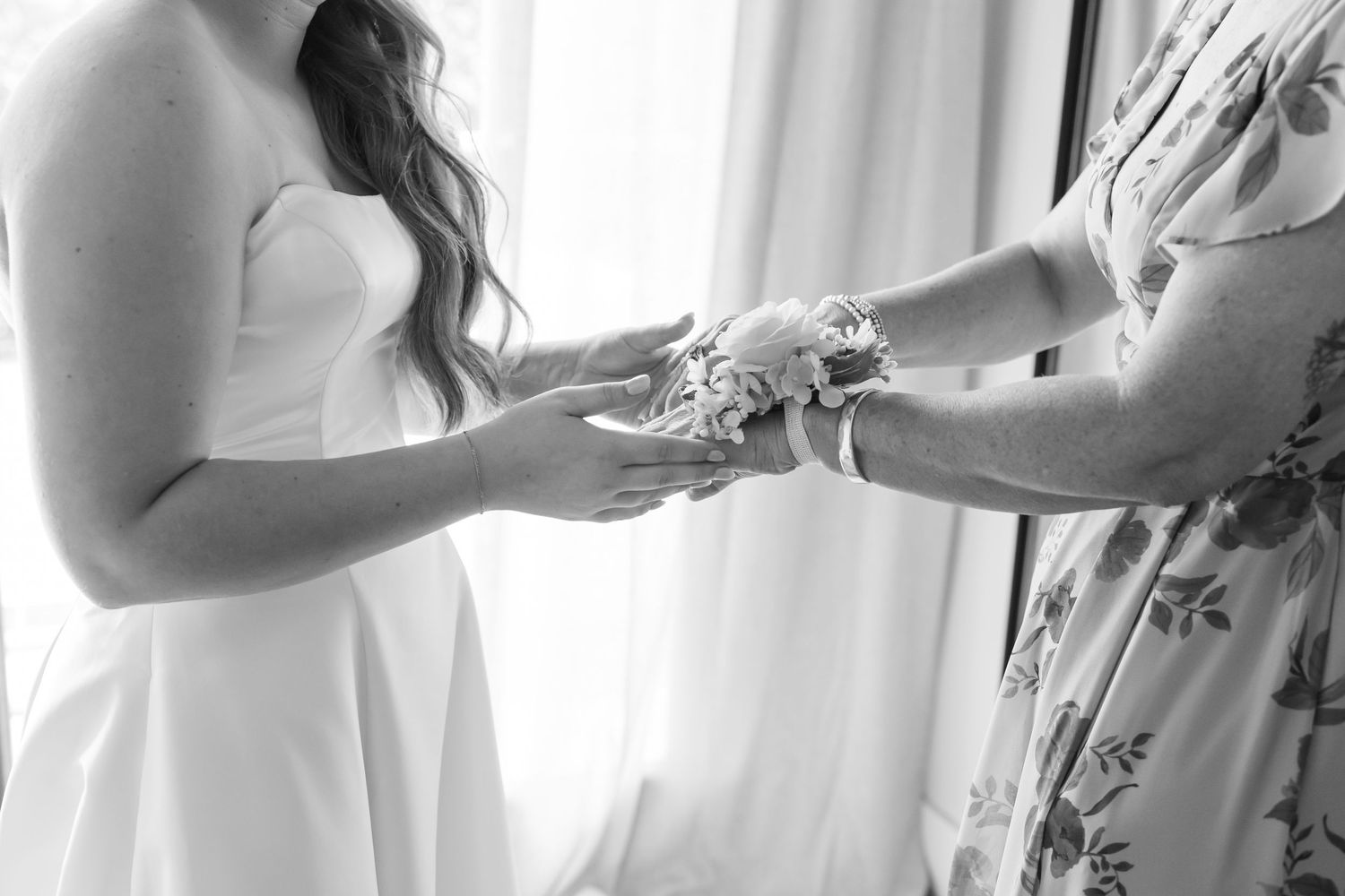Black and white photo of a bride giving a floral wrist corsage to her mother near a bright window in oklahoma.