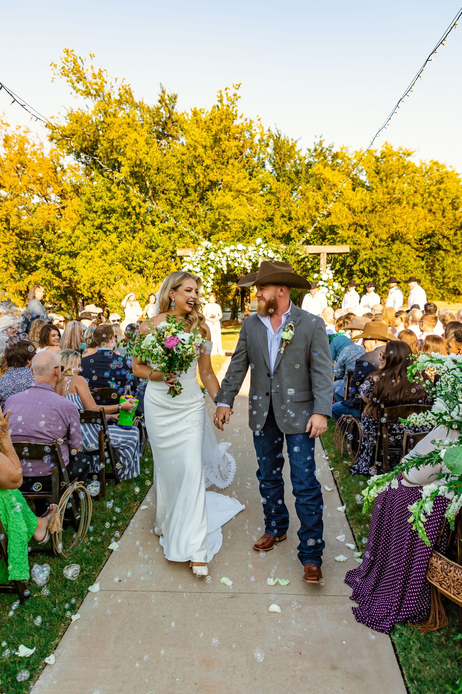 Bride and groom walk down outdoor aisle after wedding ceremony, guests seated, lush trees and floral arch backdrop at rustic rose ranch, ok