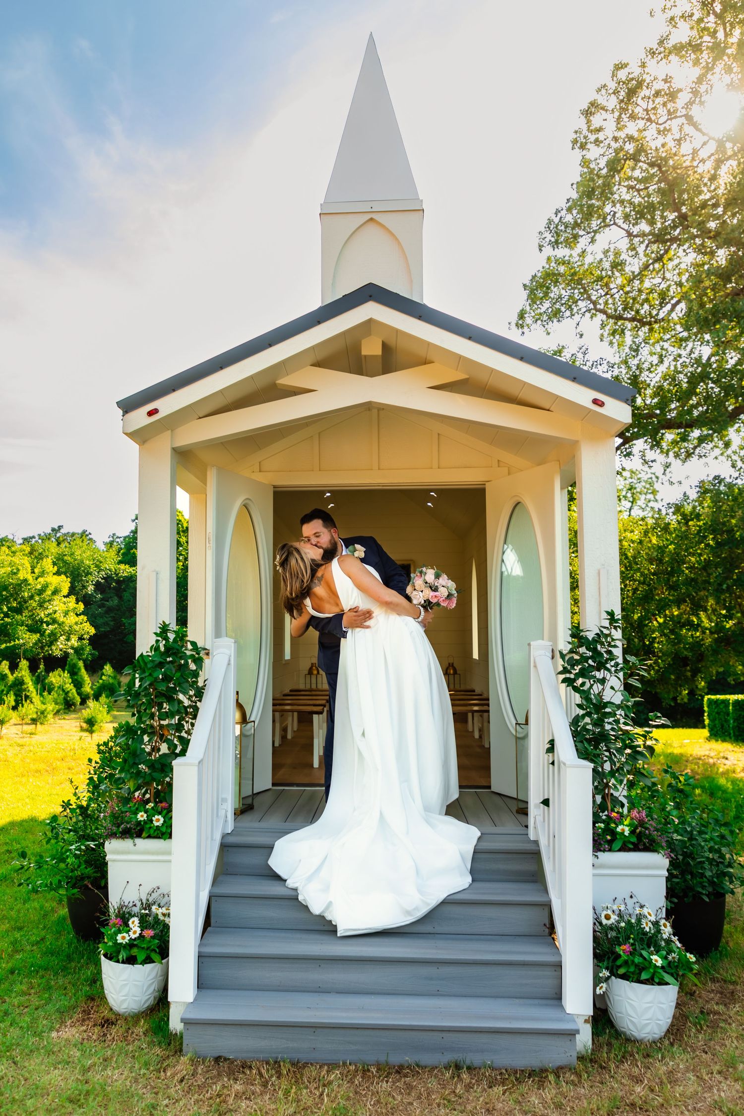 Bride and groom sharing a romantic kiss on the steps of a charming white chapel surrounded by lush greenery at wells Christmas tree farm