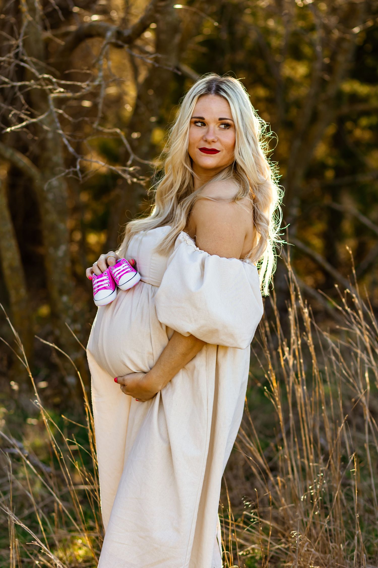 Pregnant woman in white off-shoulder dress holding pink baby sneakers outdoors in golden autumn light. maternity photo shoot in Oklahoma