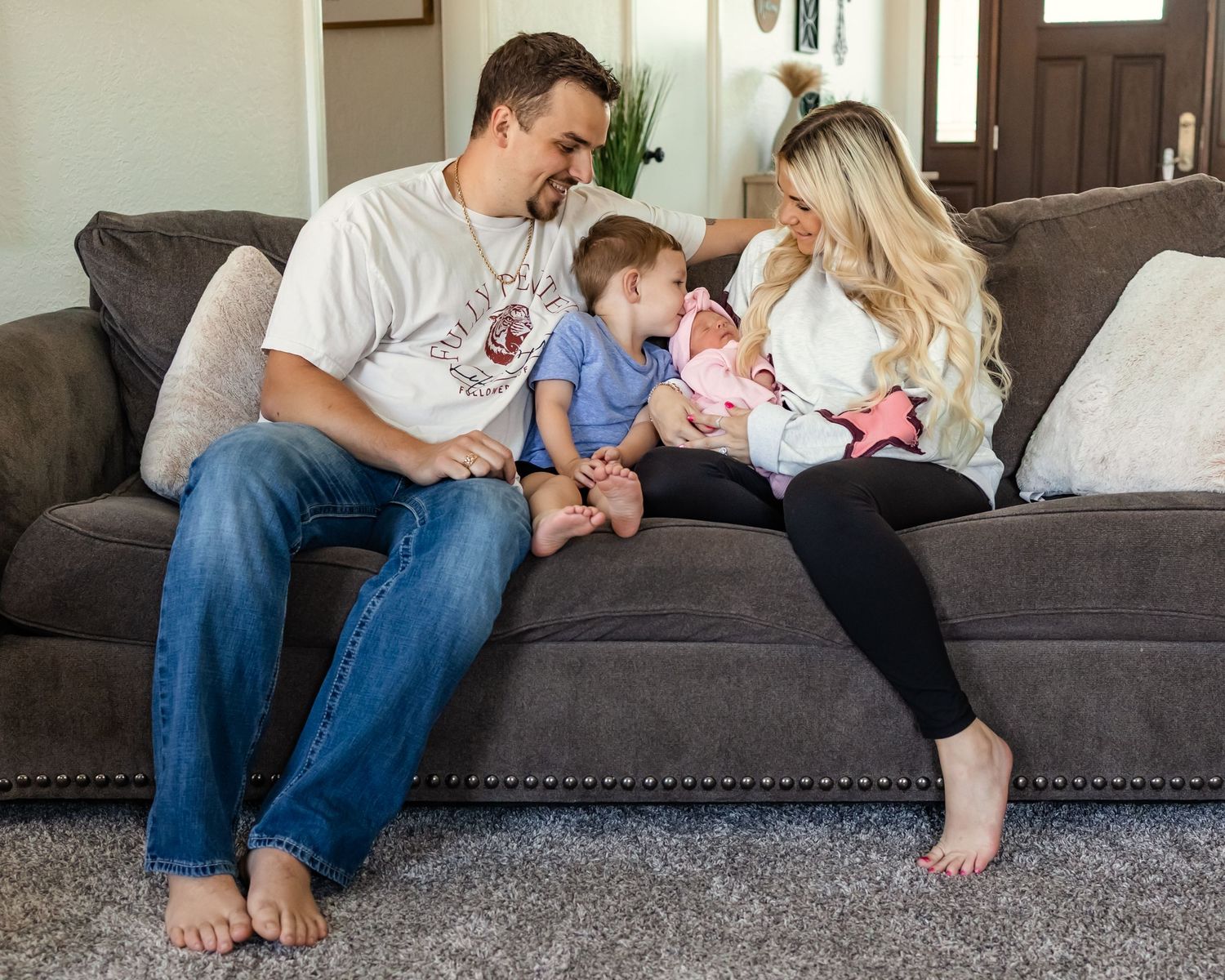 Young family with toddler and newborn baby sitting together on a gray couch in a cozy living room in Oklahoma family photography
