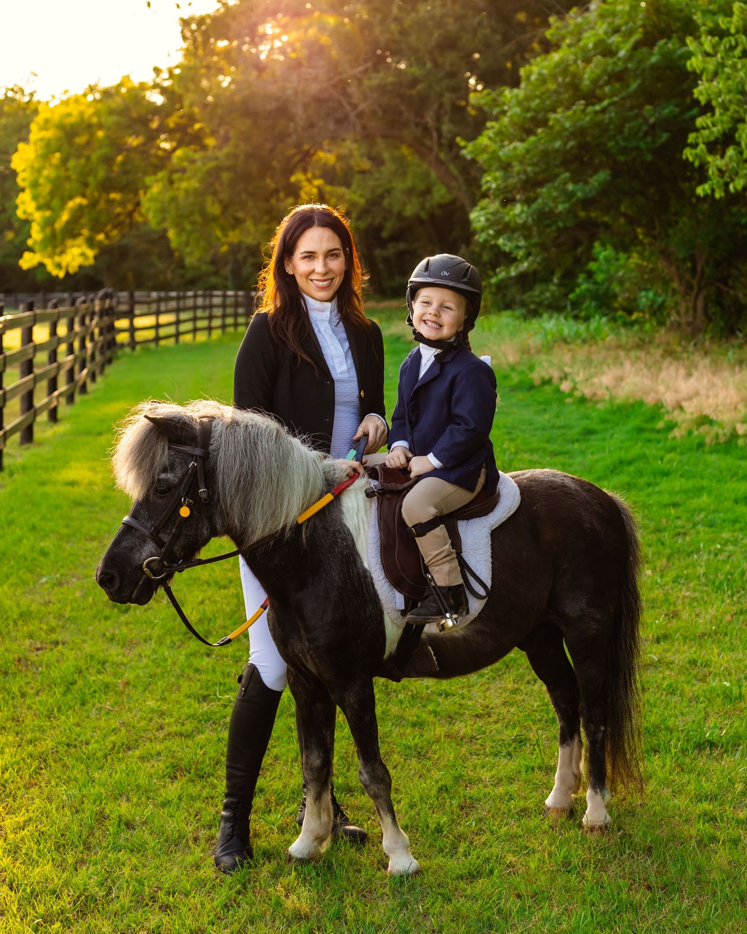 Woman and child in equestrian attire posing with a black and white pony on a sunny green paddock in Oklahoma family photography session