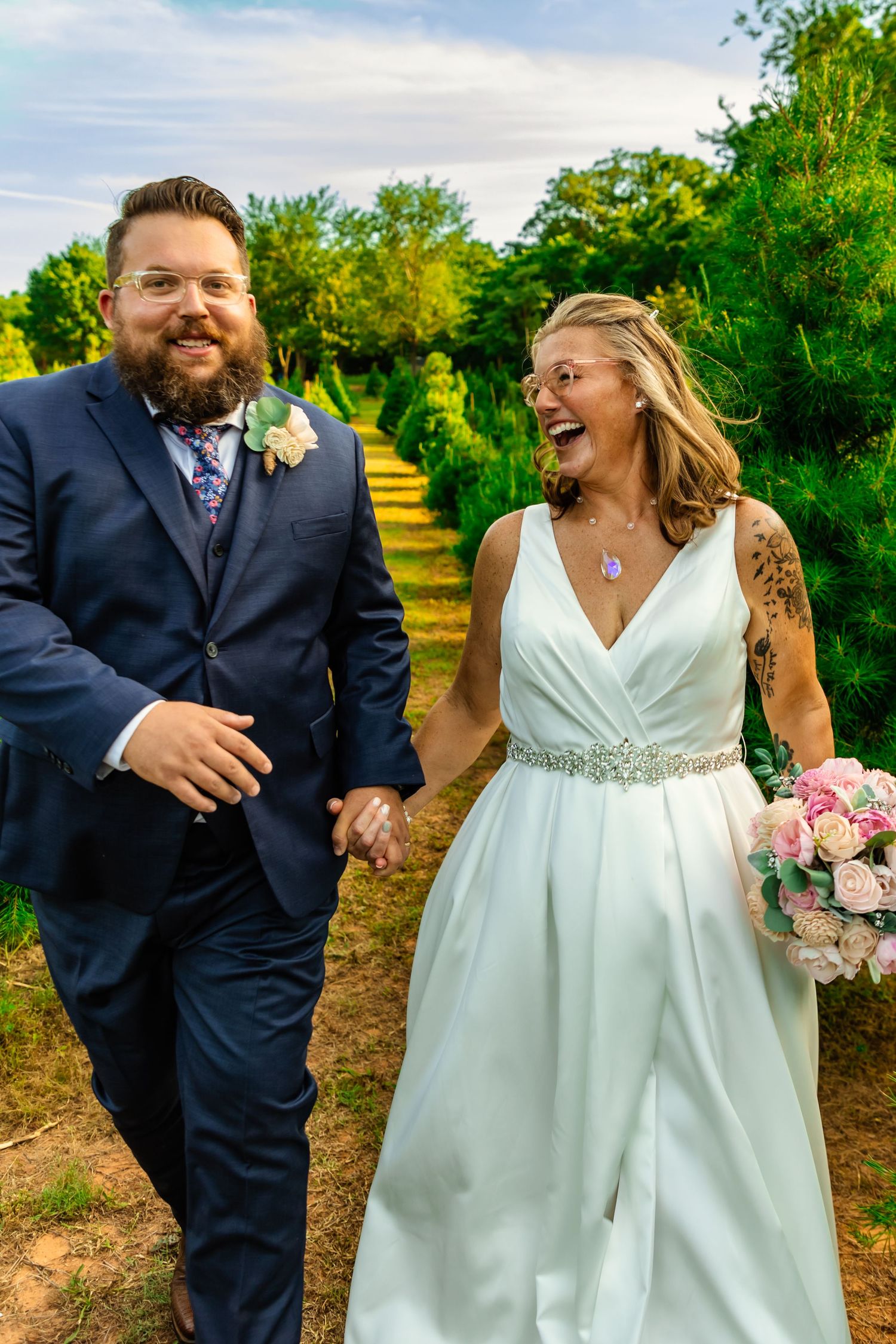 Smiling bride and groom walk hand-in-hand through lush greenery at wells Christmas tree farm. hitched traveling wedding chapel. Oklahoma