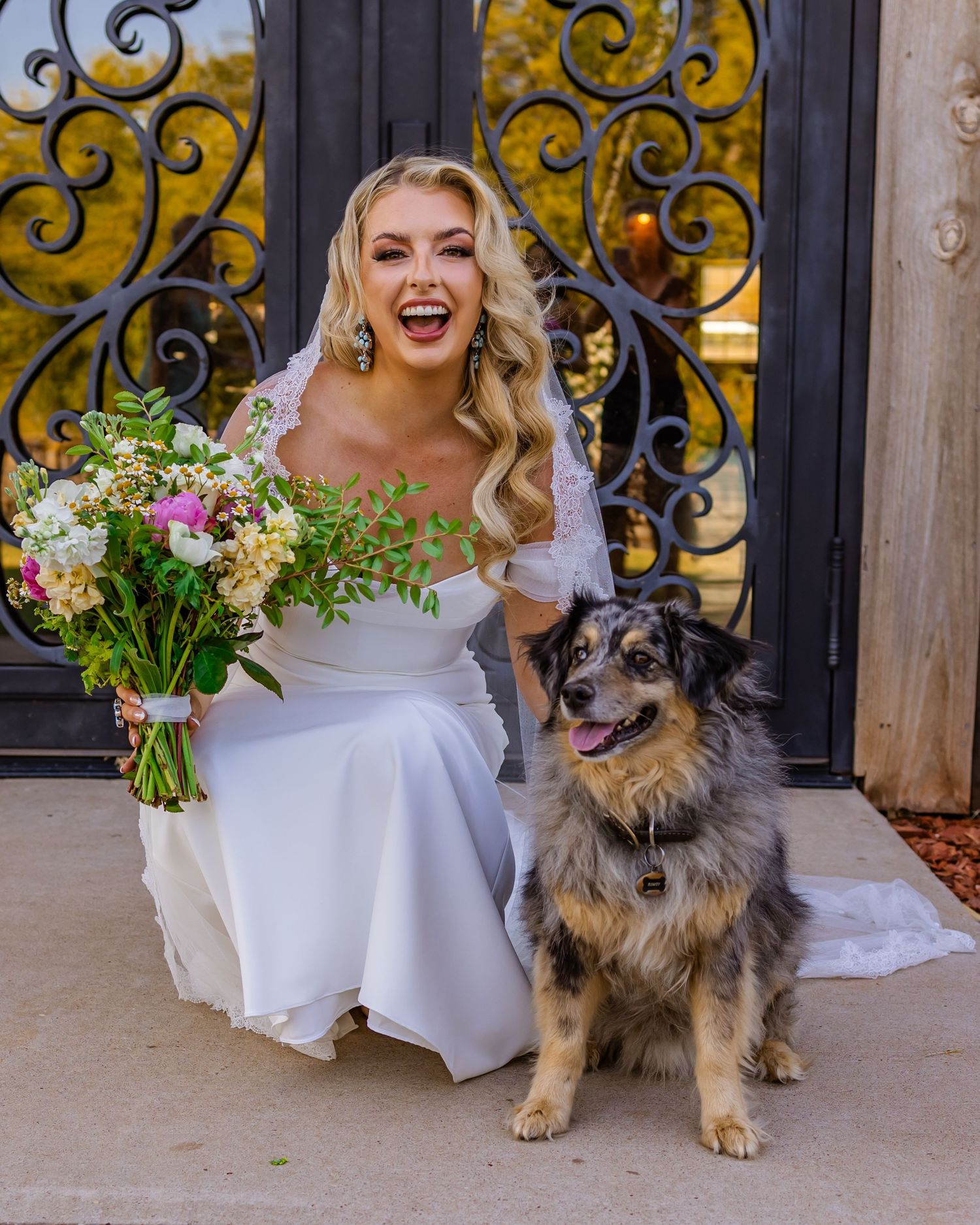 Smiling bride in white gown holding colorful bouquet poses with a Australian Shepherd dog in front of ornate iron gates at rustic rose ranch