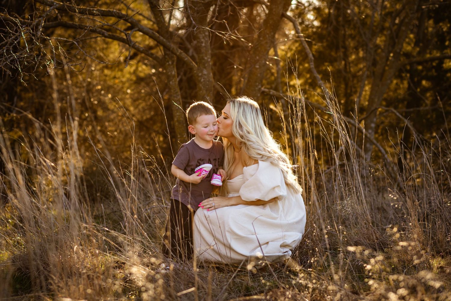 A mother kisses her young son outdoors in golden autumn light among tall grass and trees in Oklahoma maternity session.
