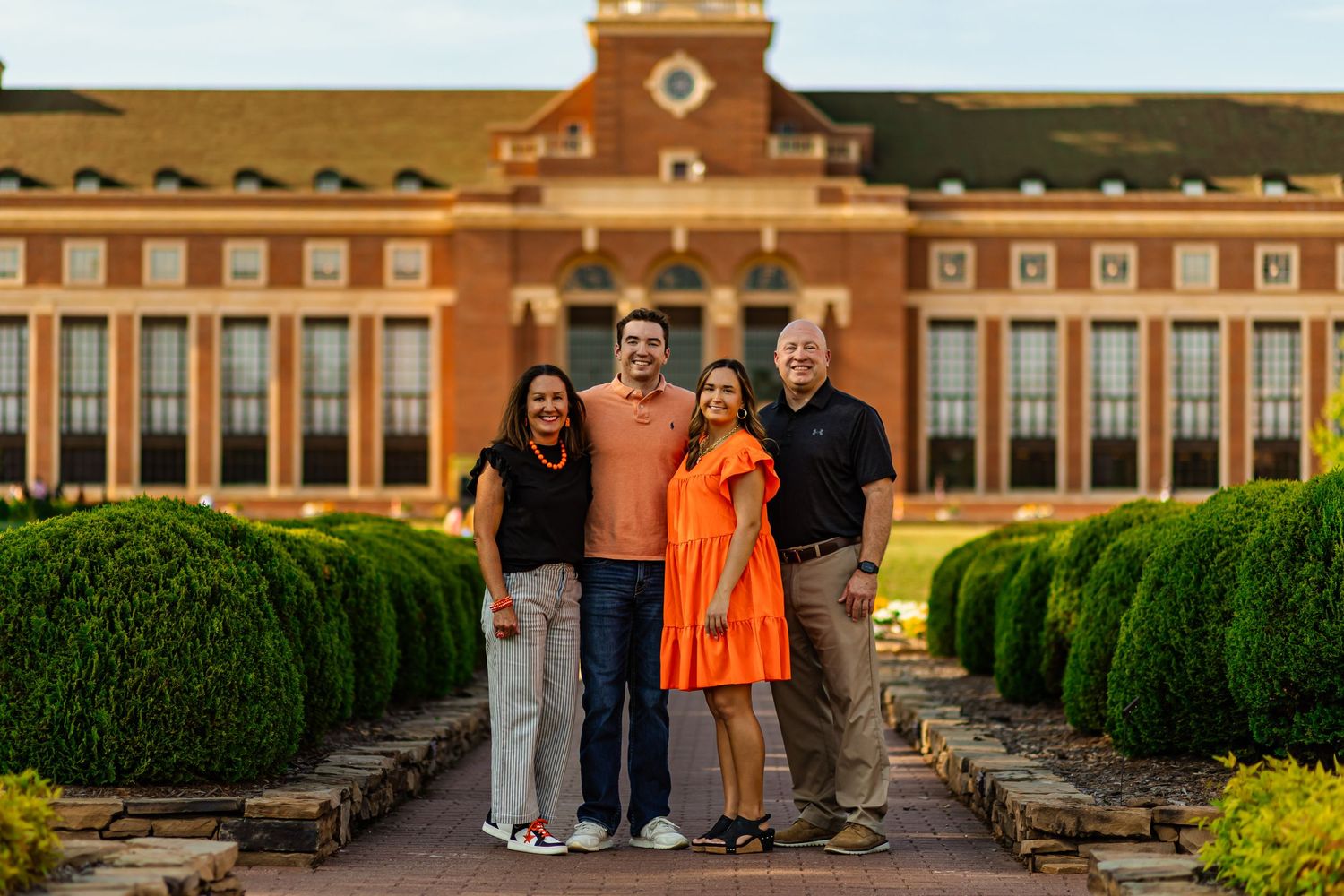 Four people smiling together outdoors in front of a red brick Oklahoma state university building surrounded by manicured hedges.