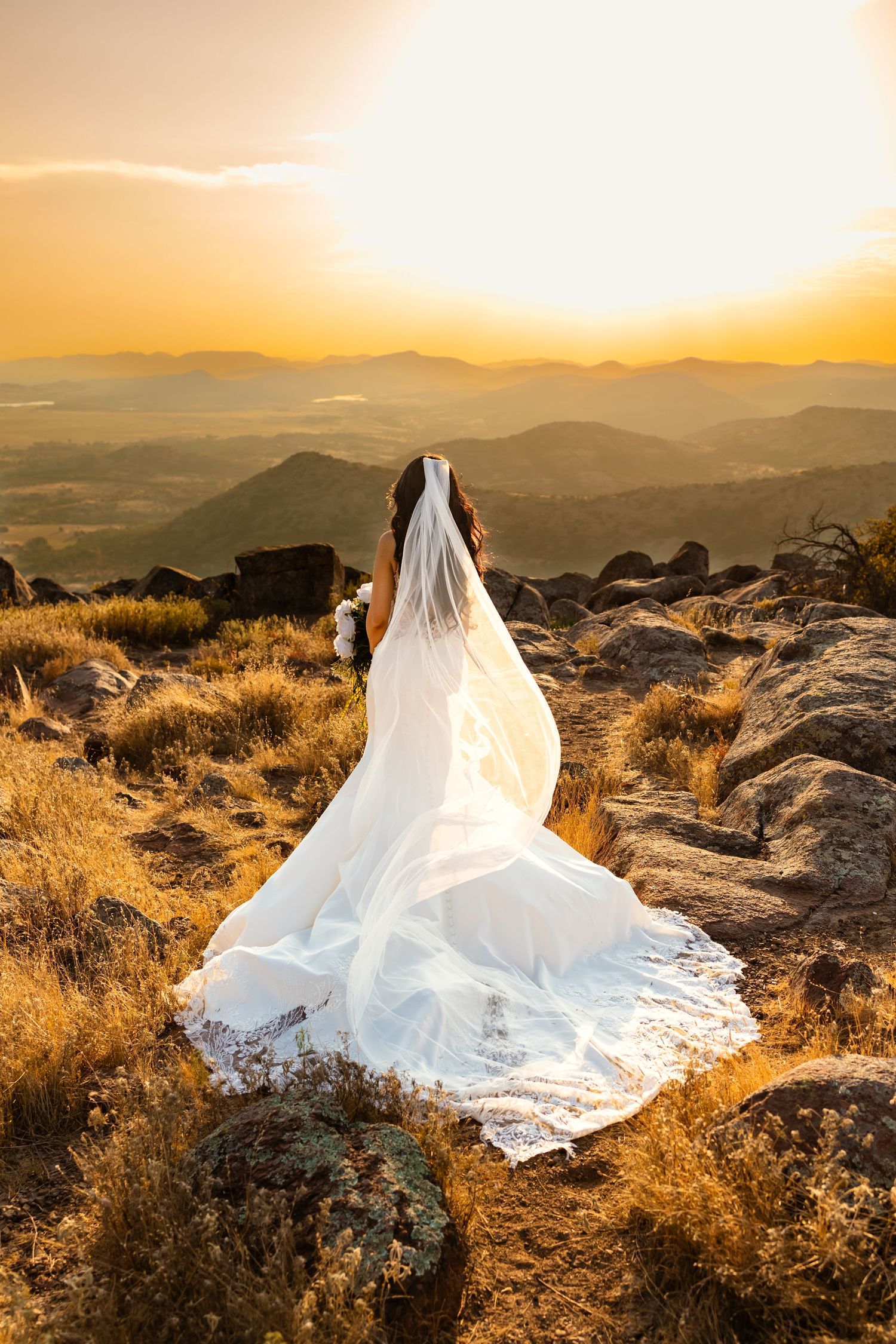 Bride in white gown and veil stands on rocky mountaintop in Oklahoma overlooking golden sunset and sweeping mountain valley views.