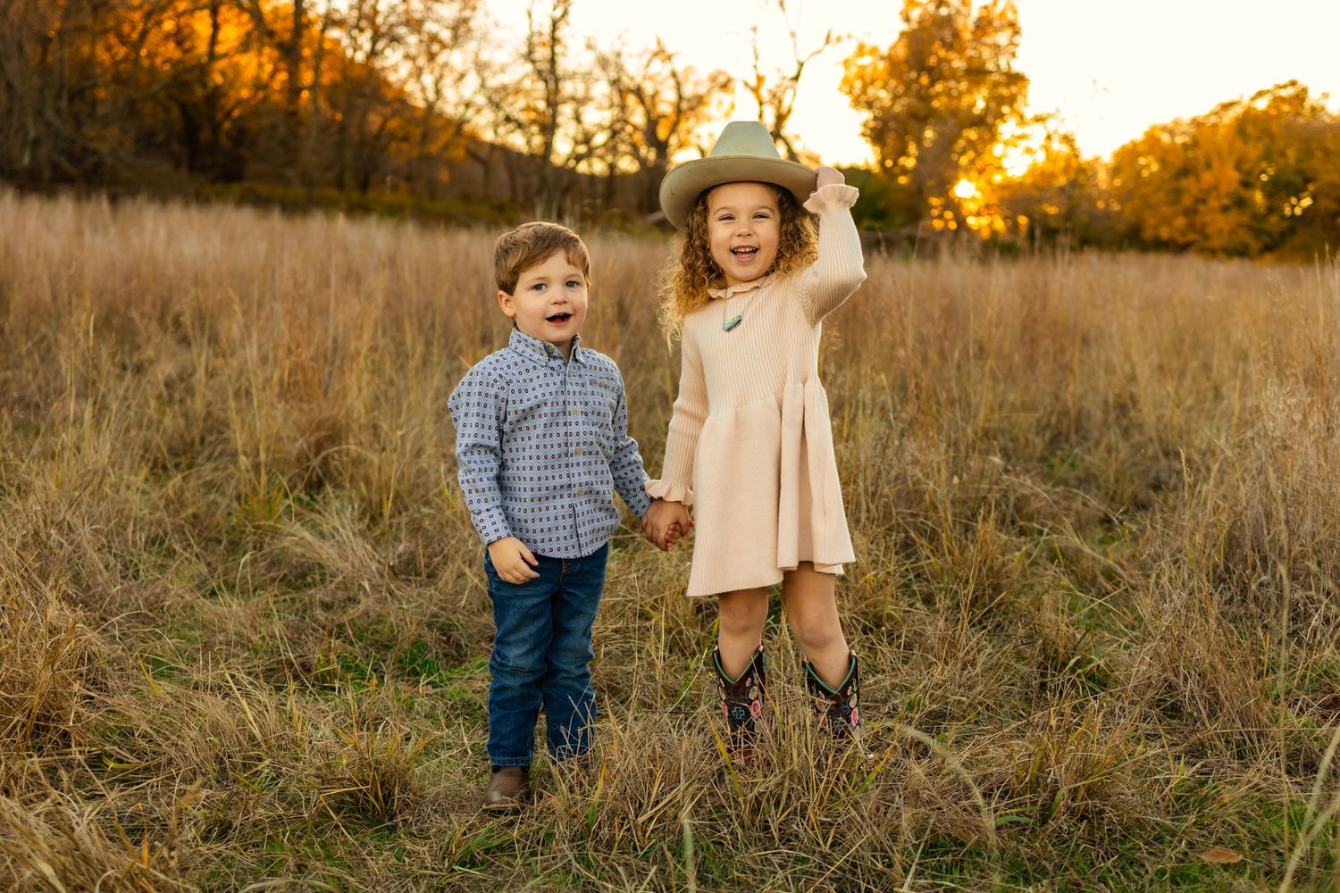 Two young children holding hands in a golden field at sunset, boy in plaid shirt and girl in dress with cowboy hat outdoor Oklahoma family