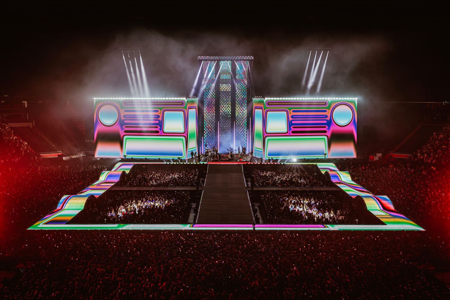 Spectacular concert stage with colorful neon lights, LED screens, and pyrotechnics dazzling a packed arena crowd at night.