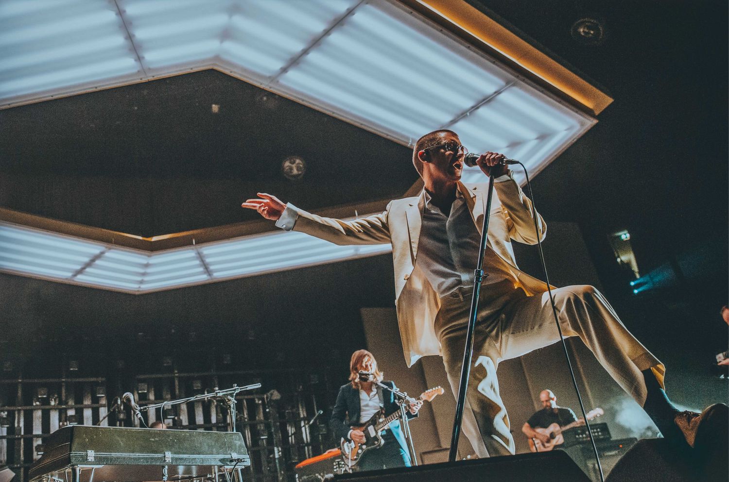 Indoor concert venue with Arctic Monkeys on stage under bright overhead lighting, directed by Ant Barrett (RESOLVE FILM)