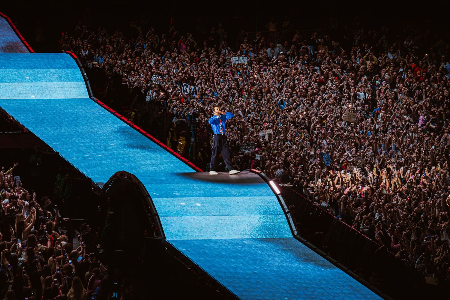 A performer stands on a illuminated blue stage runway before a massive crowd at a large indoor concert venue.