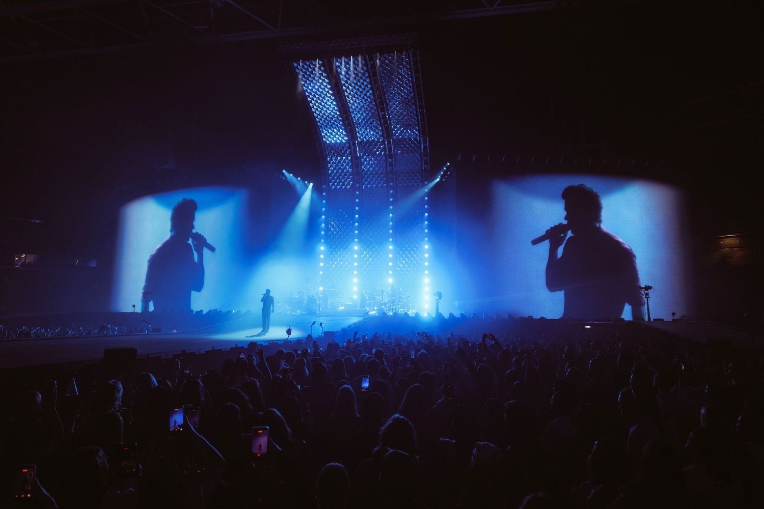 Live concert with two performers on stage, dramatic blue lighting, large crowd, and giant projection screens.