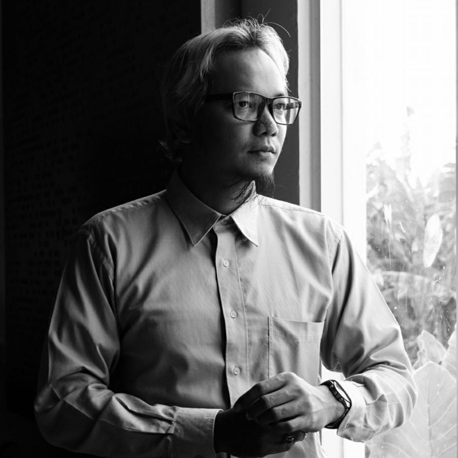 Professional black and white portrait of person wearing glasses and collared shirt looking pensively by window.