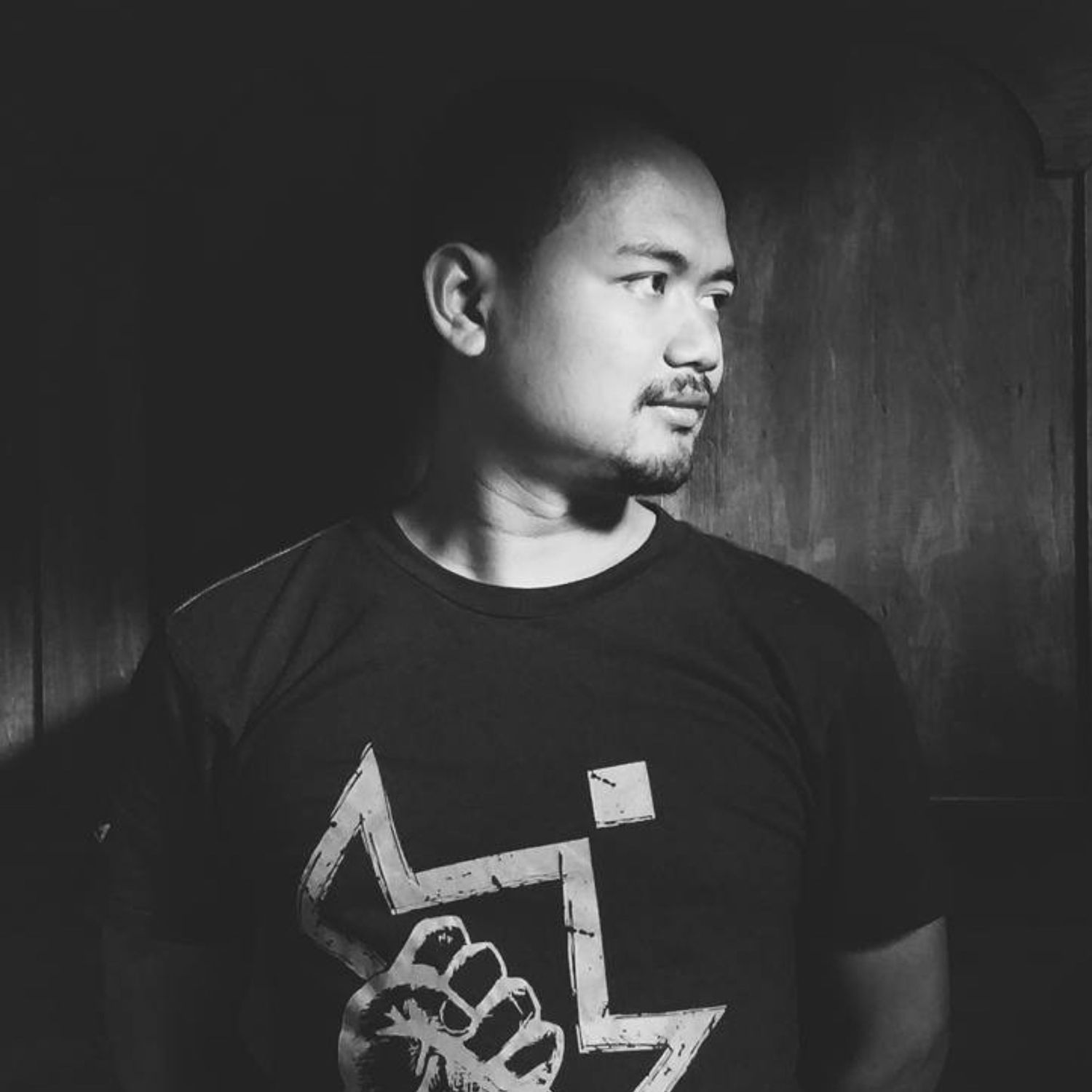 Dramatic black and white side profile portrait of person wearing graphic t-shirt against dark background.