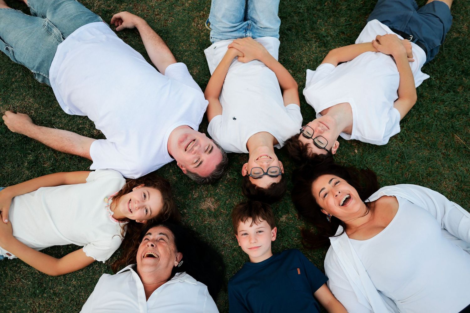 Group of people laying in grass wearing white shirts arranged in a circle formation while laughing together.