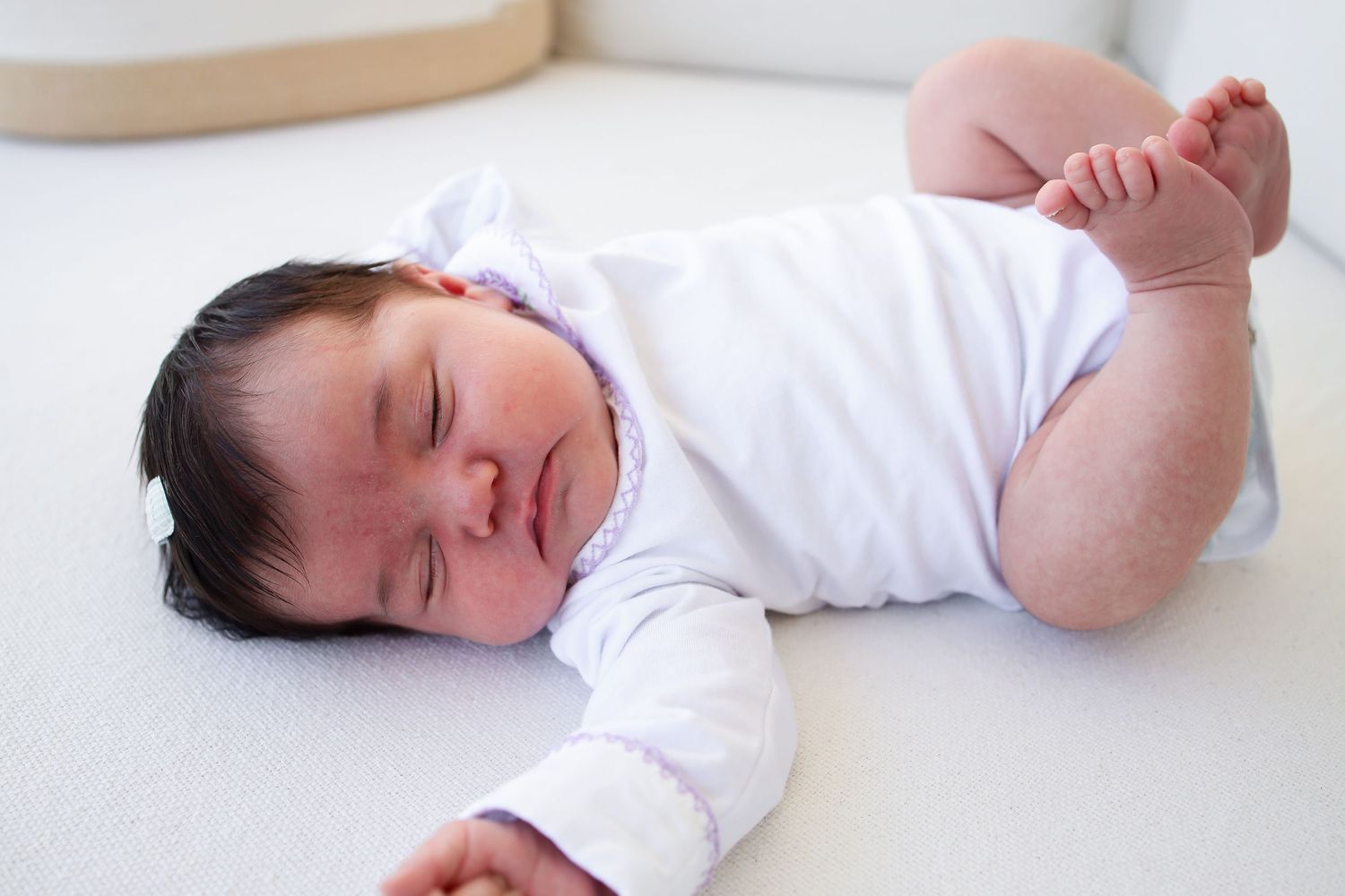 A sleeping newborn baby wearing a white onesie lies peacefully on white bedding.
