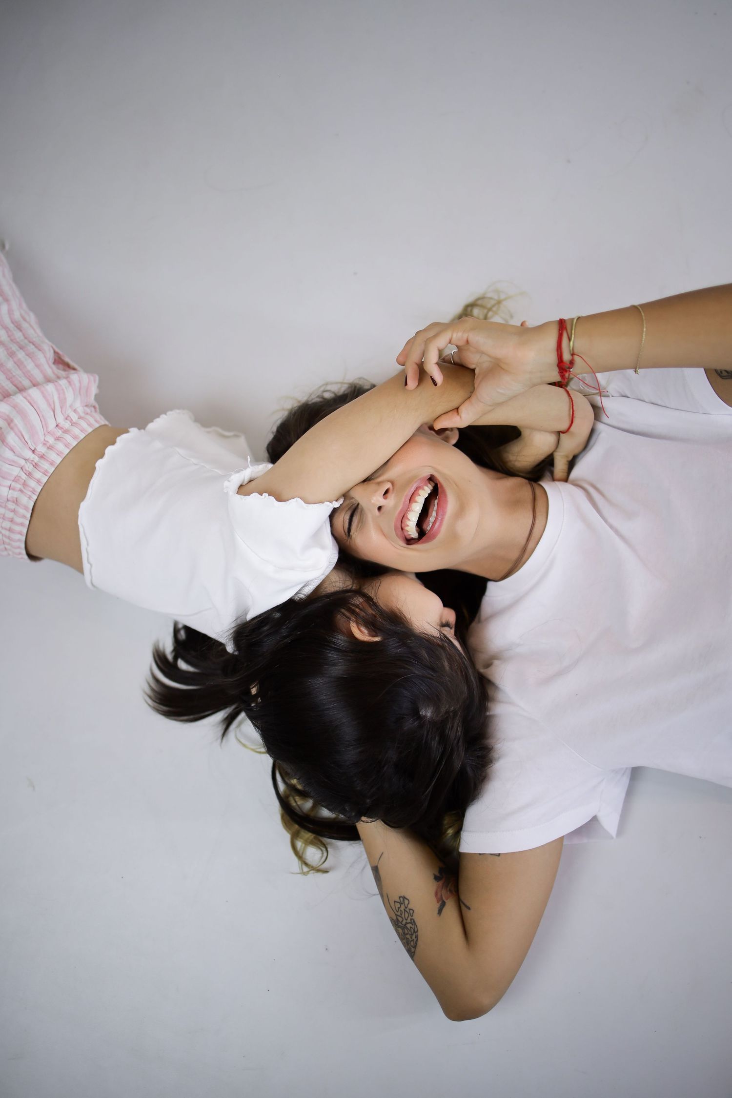 Two young women laughing and lying on a white floor, one covering her face while wearing white tops.