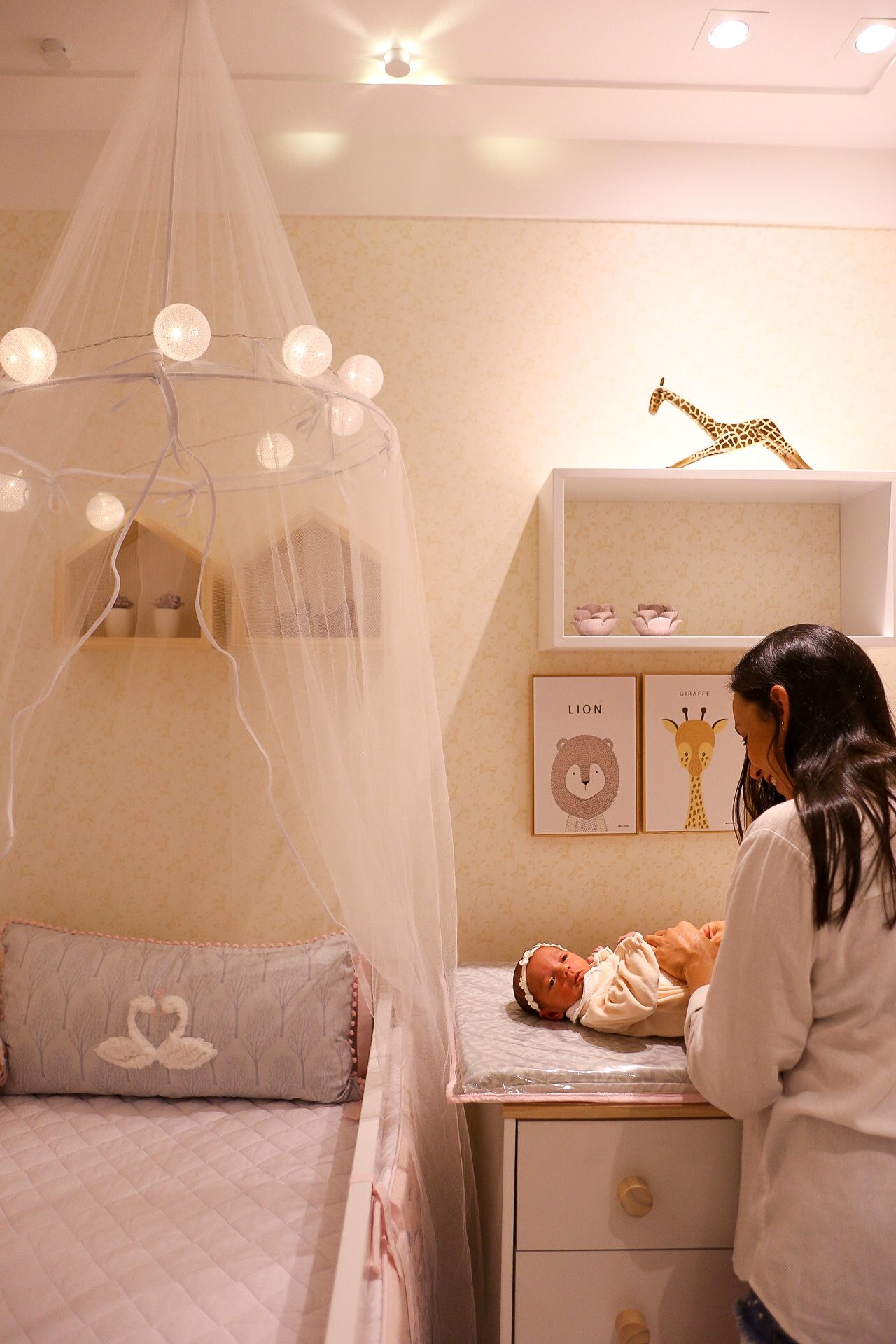 A cozy nursery corner with sheer canopy, string lights, and gold giraffe decor features white and beige tones.