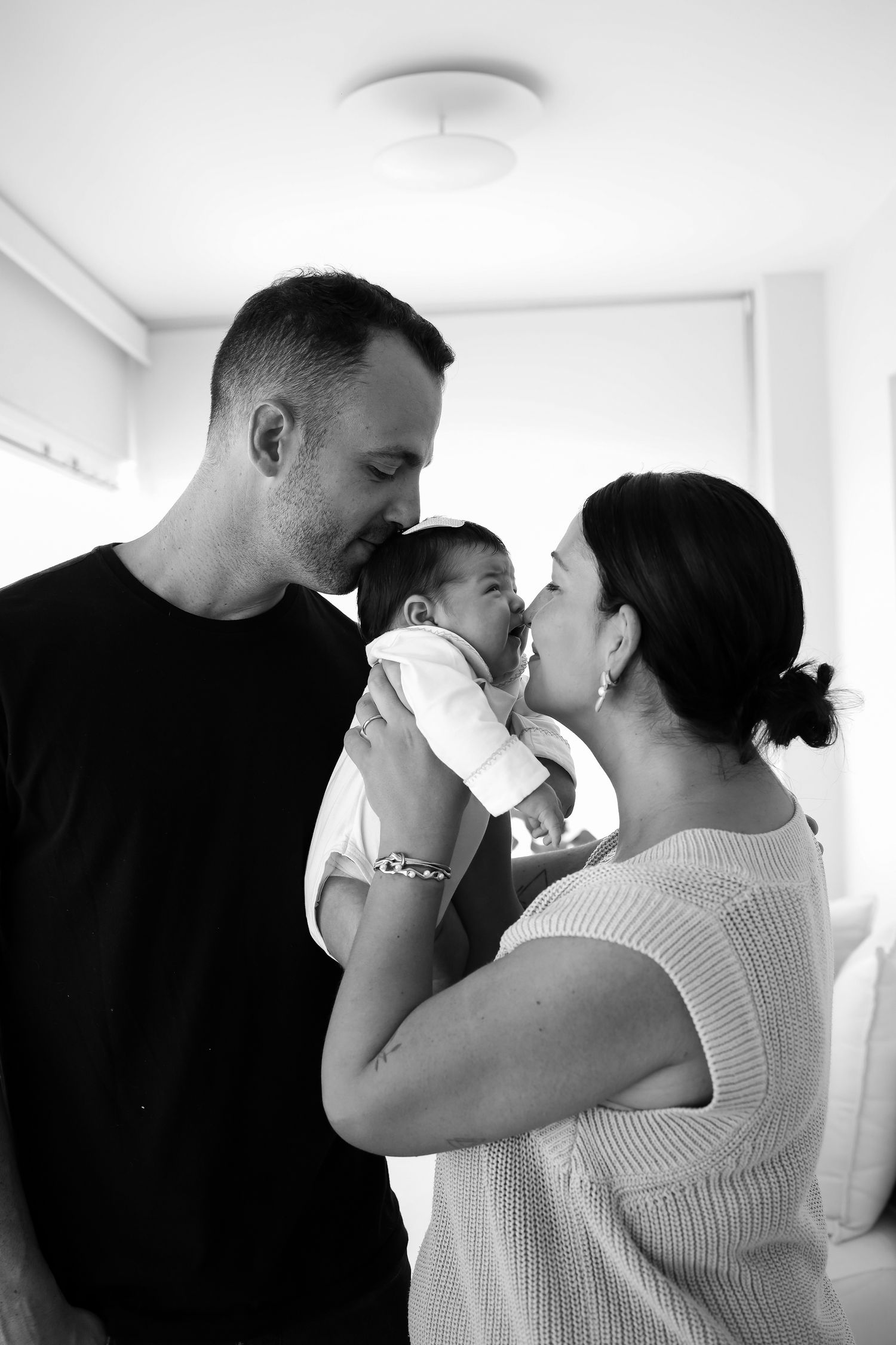 A tender black and white family moment with parents sharing a gentle kiss with their baby between them.