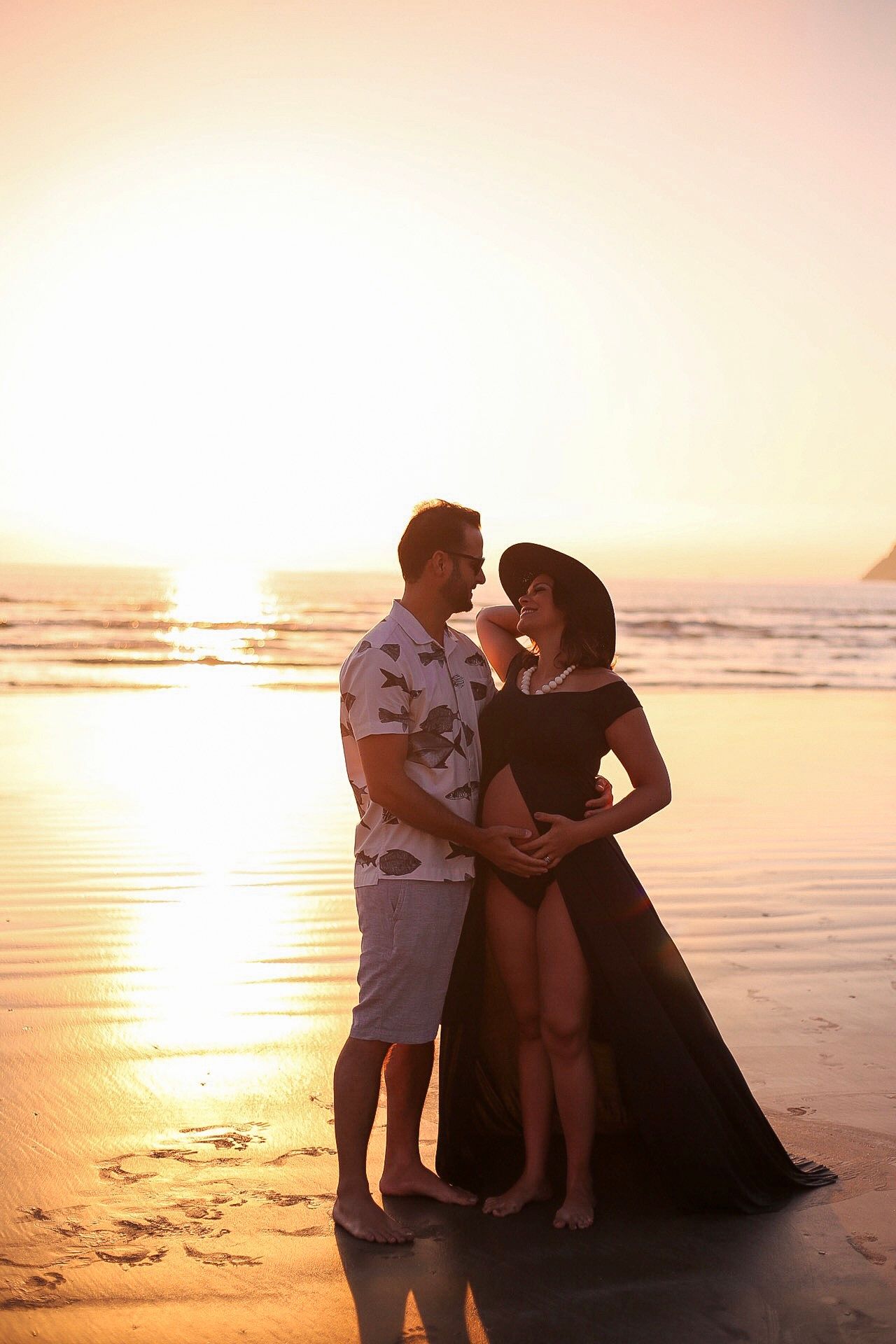 A couple embrace on a beach at sunset while standing in golden light reflecting off the ocean and wet sand.