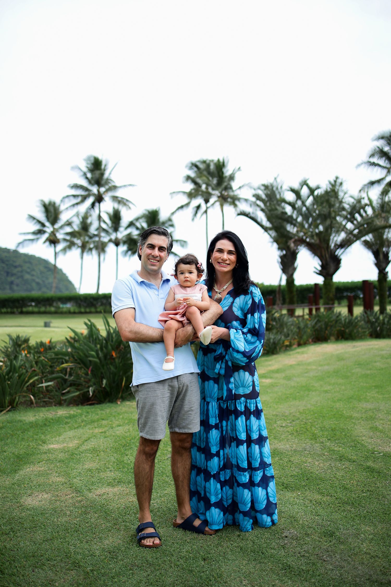 A family poses outdoors on a grassy lawn with tropical palm trees and mountains in the background.