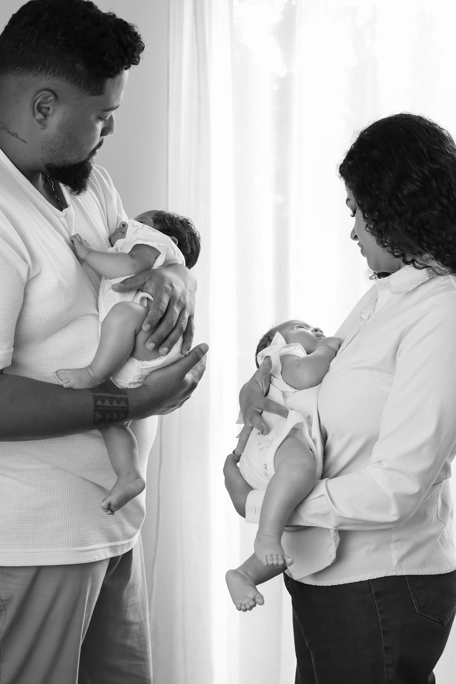 Black and white photo of parents in white tops holding twin babies near a white curtained window.
