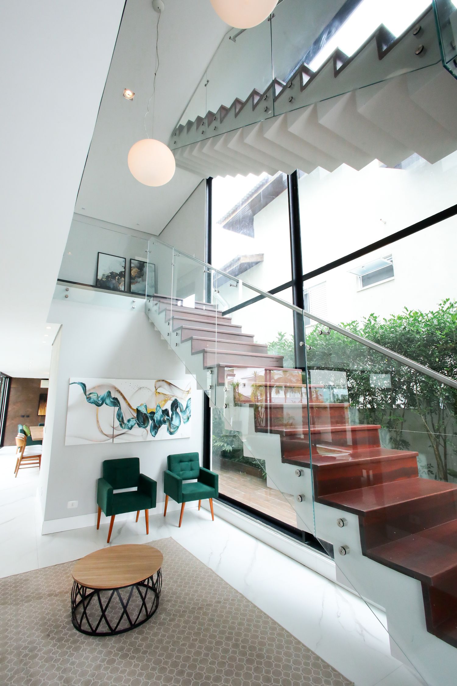 Modern interior staircase with glass railings, green accent chairs, and floor-to-ceiling windows in a contemporary living space.