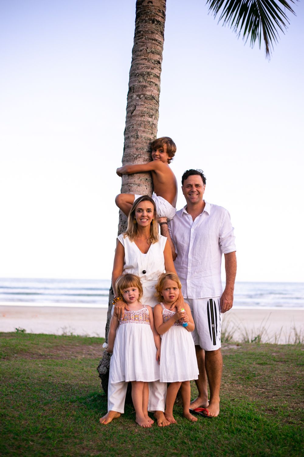 Family poses together on grassy area by beach with palm tree trunk and ocean in background during sunset.