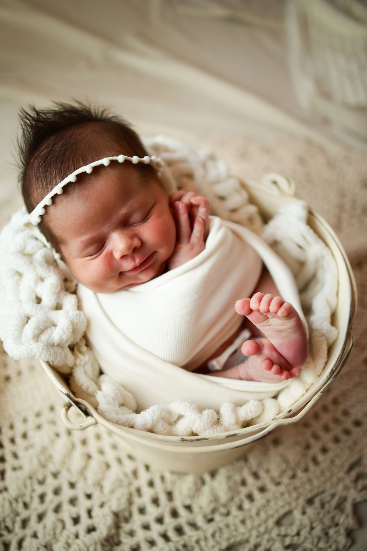 Newborn baby peacefully sleeping in a white knit basket wearing delicate headband and wrapped in cream swaddle.