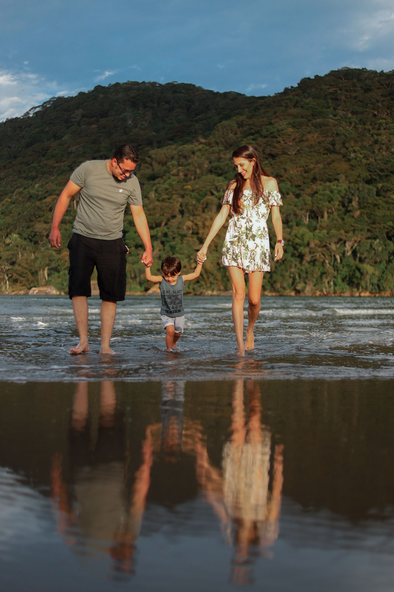 A couple bends down to play in the shallow water at the beach with mountains and forest in the background.