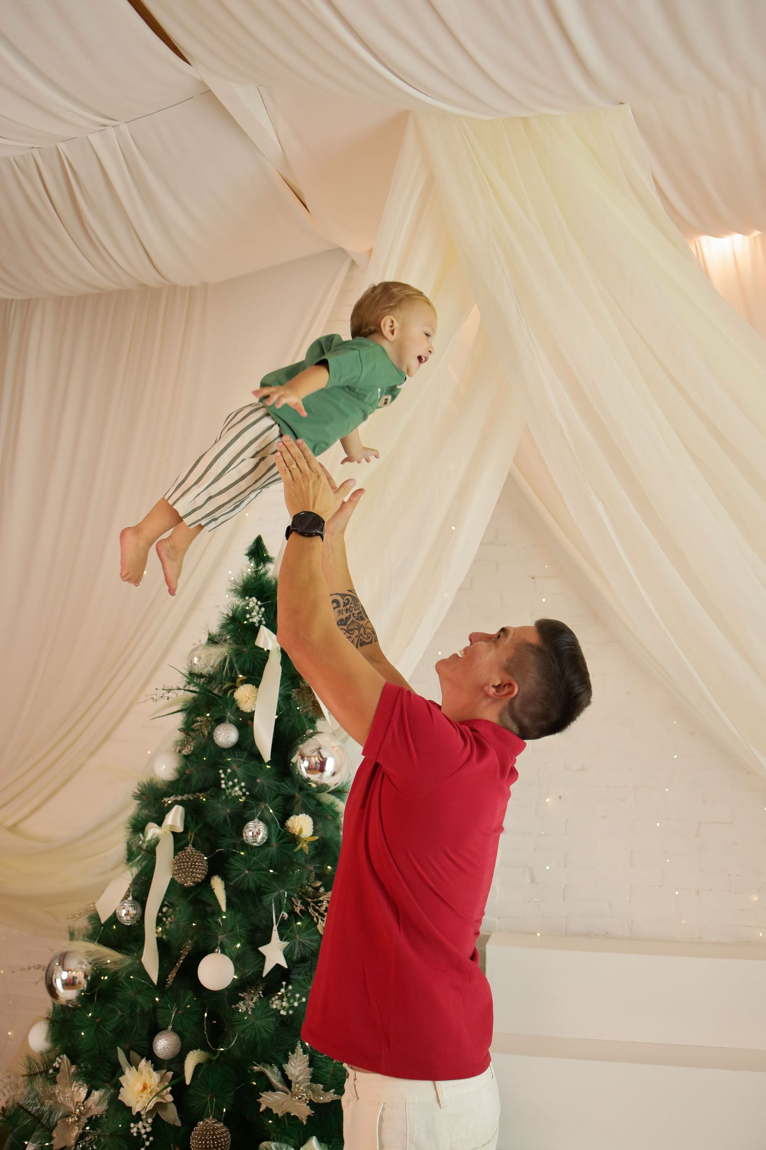 A person in a red shirt lifts a small child up to place them on top of a decorated Christmas tree in a white draped room.
