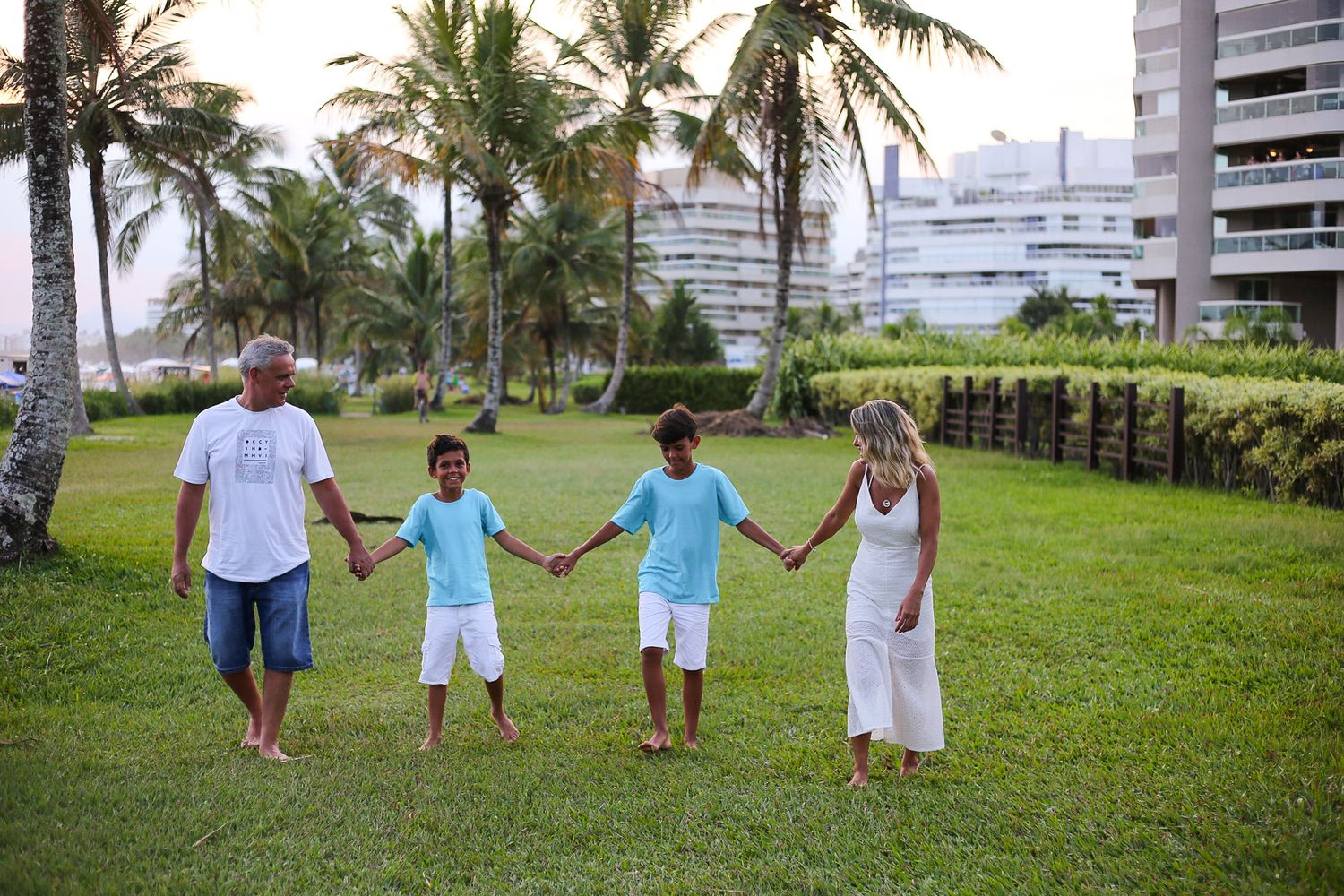 Family members holding hands while walking together on a grassy lawn with palm trees and city buildings in background.