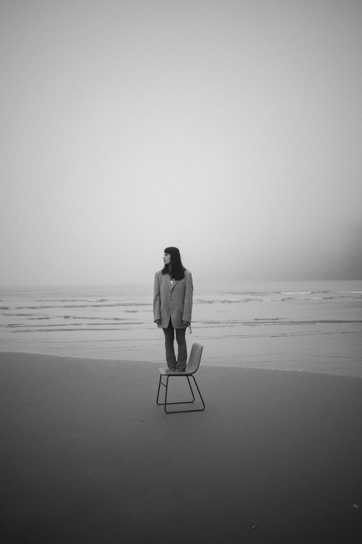 An artistic black and white photo of a person standing on a chair on an empty beach with a foggy horizon.