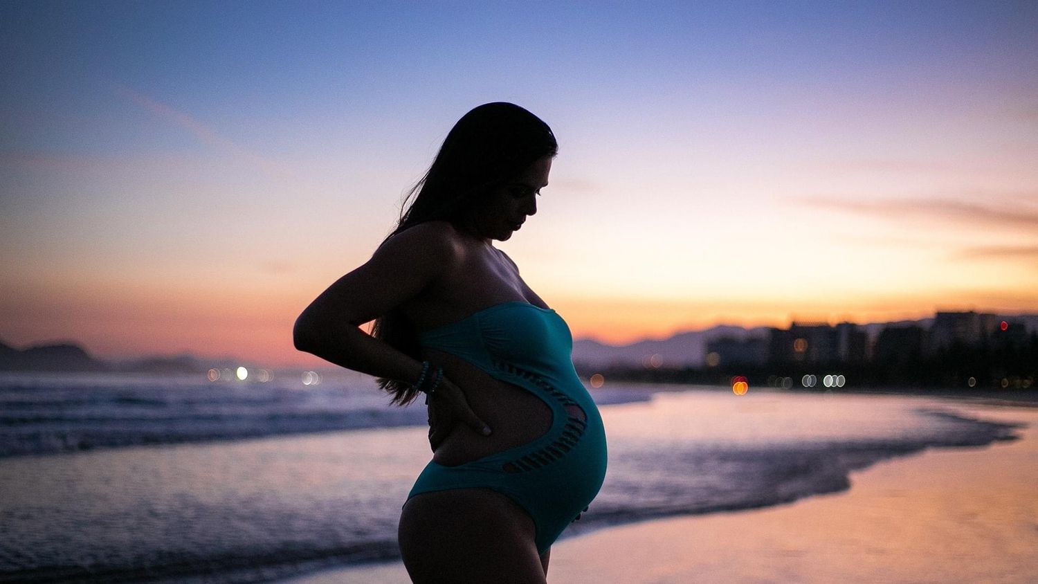 Silhouette of pregnant woman standing on beach at sunset with ocean waves and colorful sky in background.