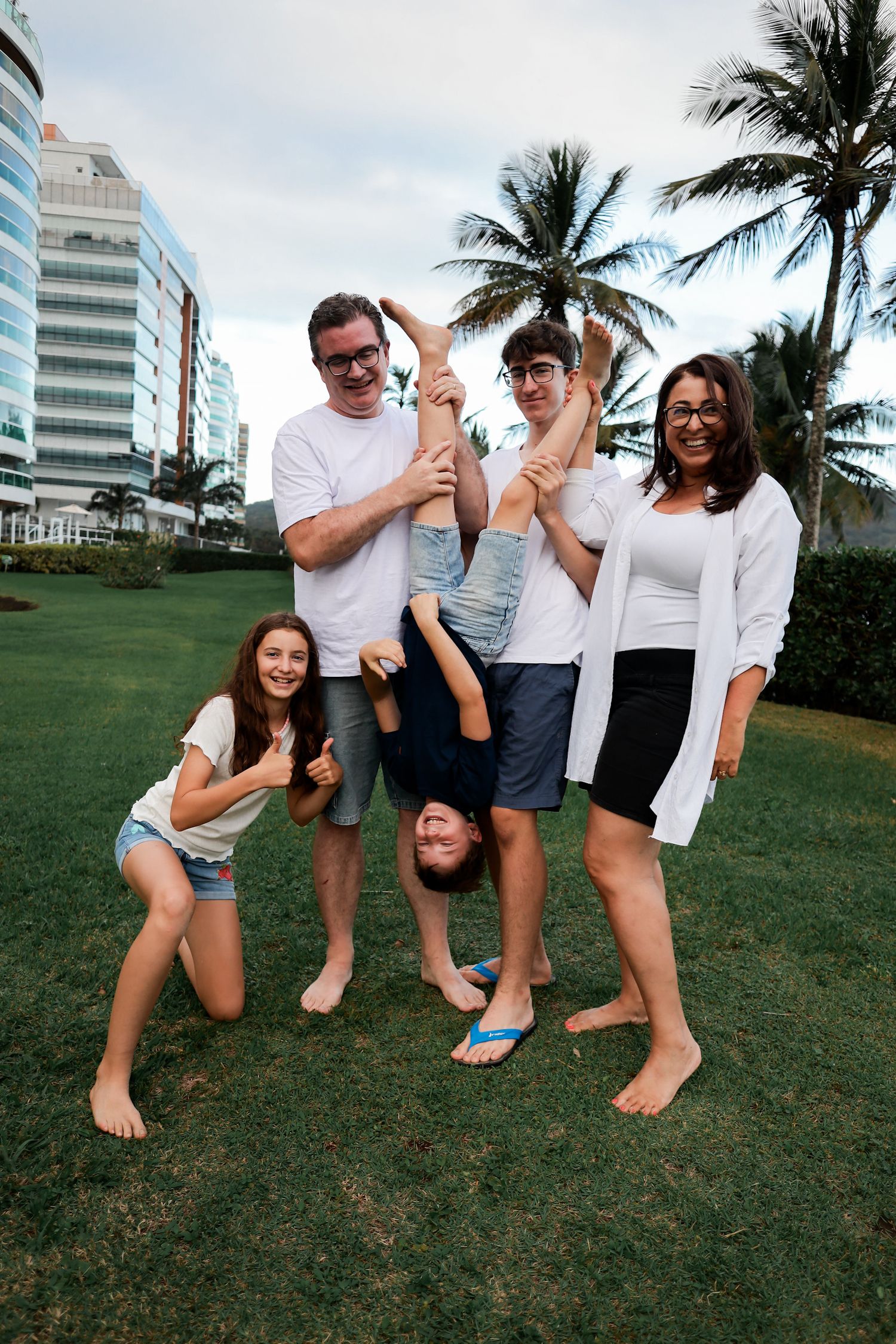 A group in white clothing poses playfully on a grassy lawn with palm trees and buildings in the background.