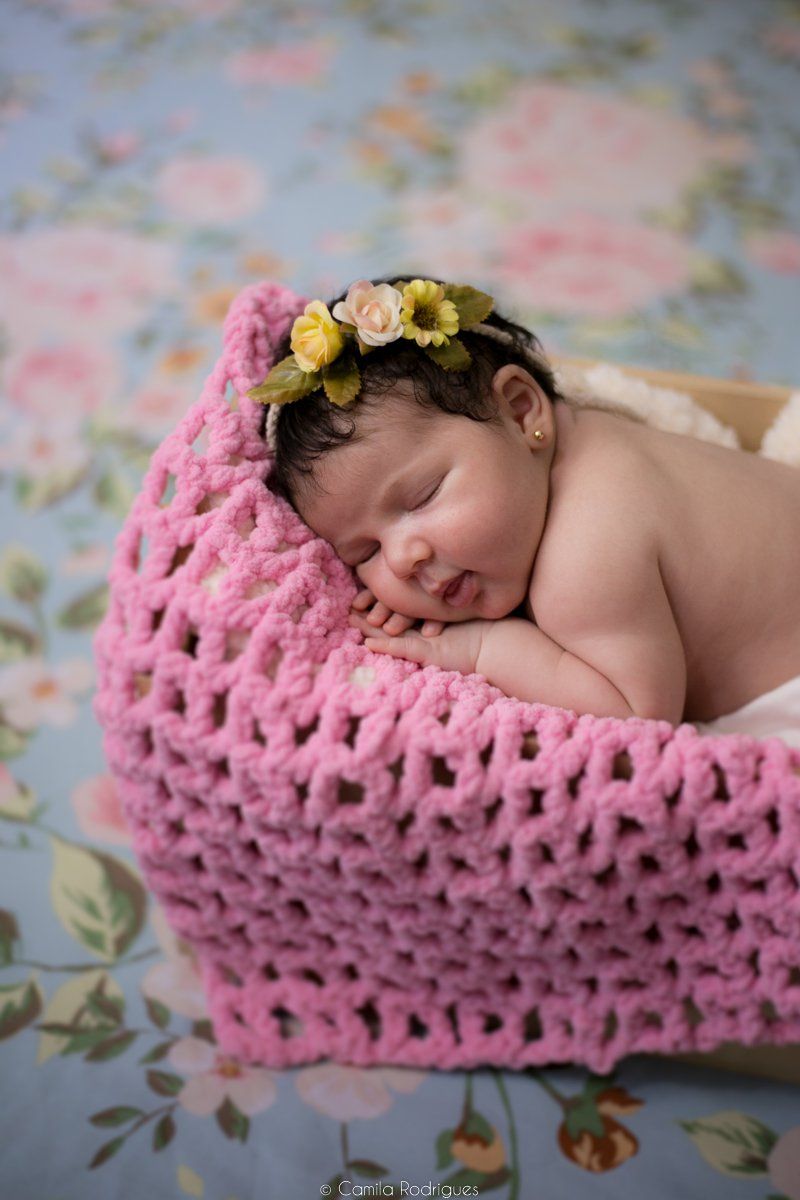 Sleeping newborn wearing yellow and pink flower crown nestled in cozy pink knit blanket on floral background.