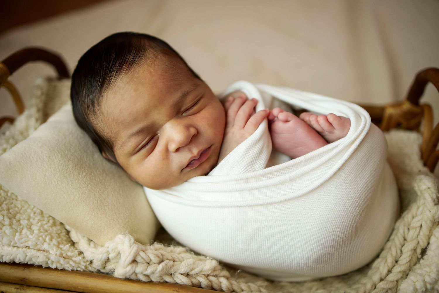 Peaceful newborn baby wrapped in white swaddle blanket sleeping on a beige blanket.