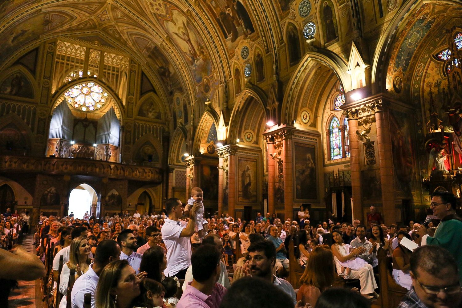 Large crowd gathers inside ornate cathedral with vaulted ceilings, stained glass windows, and golden architectural details.