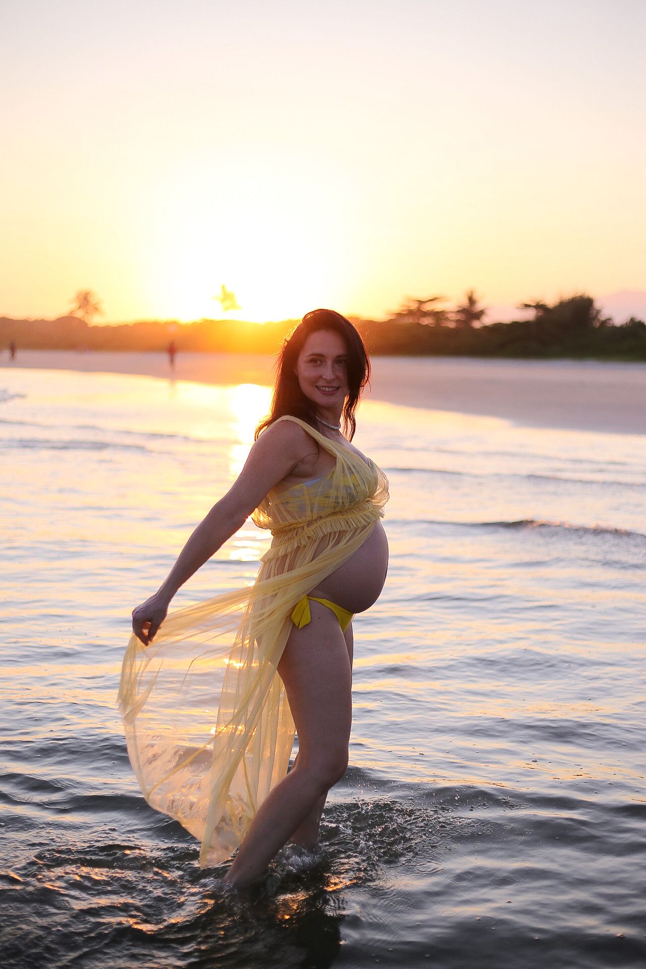 Person in flowing yellow dress stands in ocean water during dramatic beach sunset.