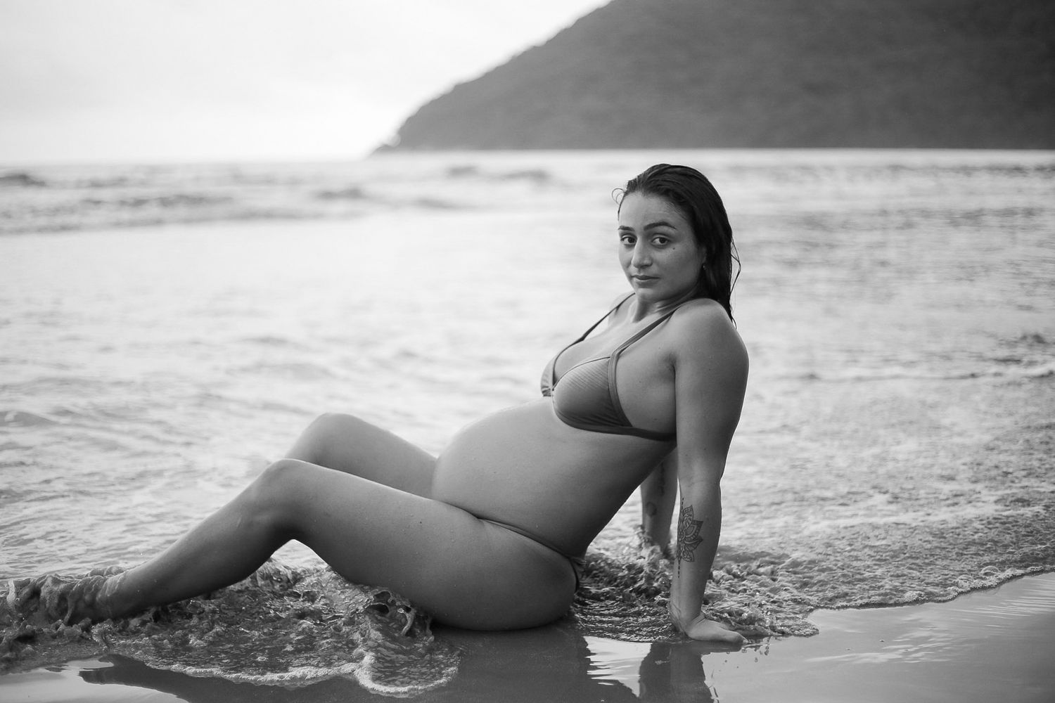 Black and white photograph of an expectant mother posing in a swimsuit on a misty beach at sunset.