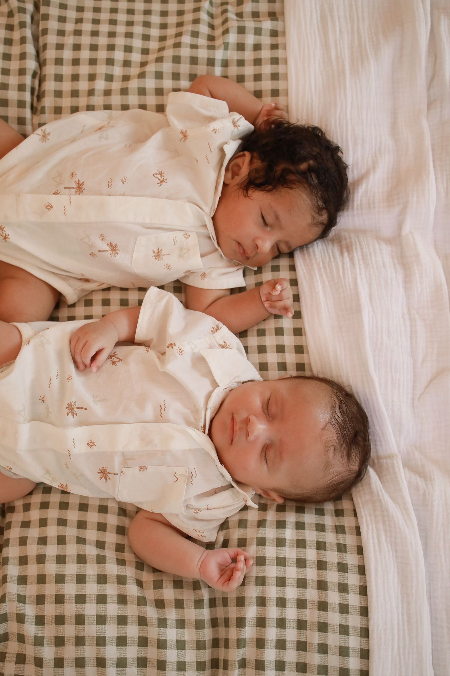 Two babies sleeping peacefully side by side on a gingham blanket wearing matching white floral outfits.