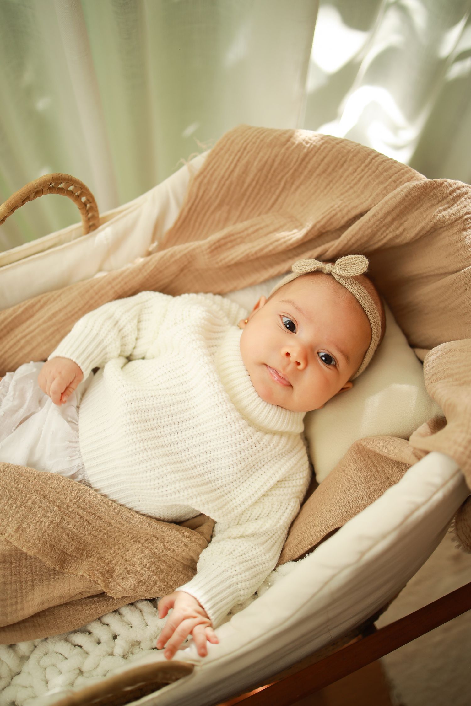 Baby wearing white knit sweater rests in cozy beige bassinet with bow headband.