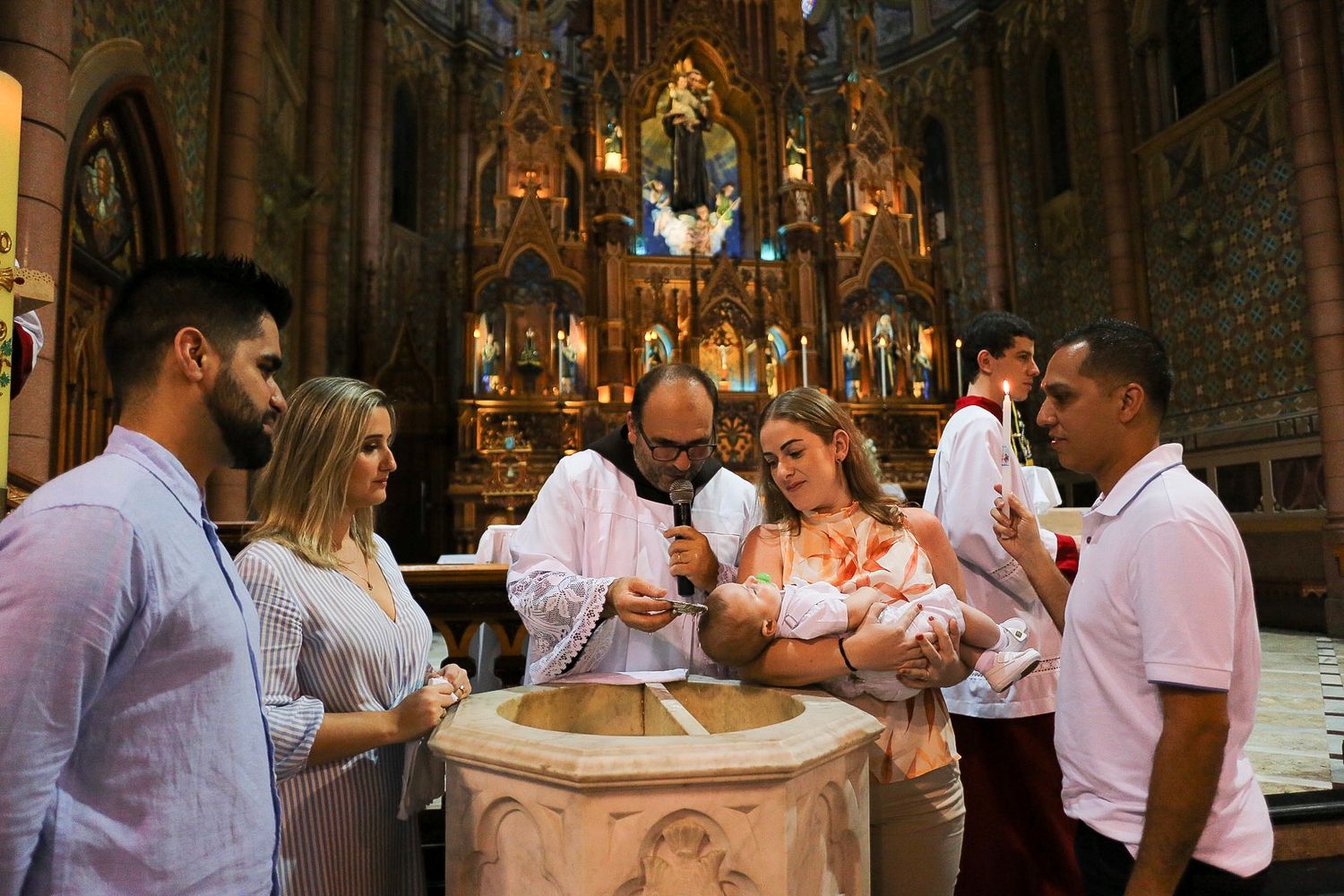 A religious baptism ceremony takes place at an ornate stone baptismal font inside a beautiful Gothic cathedral.
